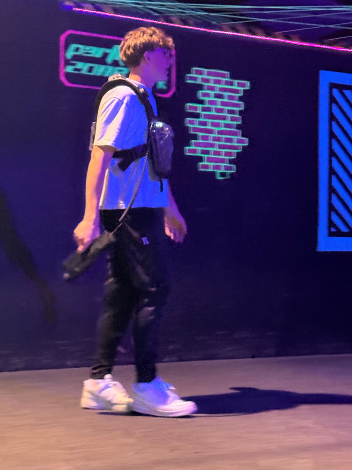 Ein junger Mann mit Lasertag-Weste und Laserpistole geht durch eine abgedunkelte Halle mit neonfarbenen Wandmustern.