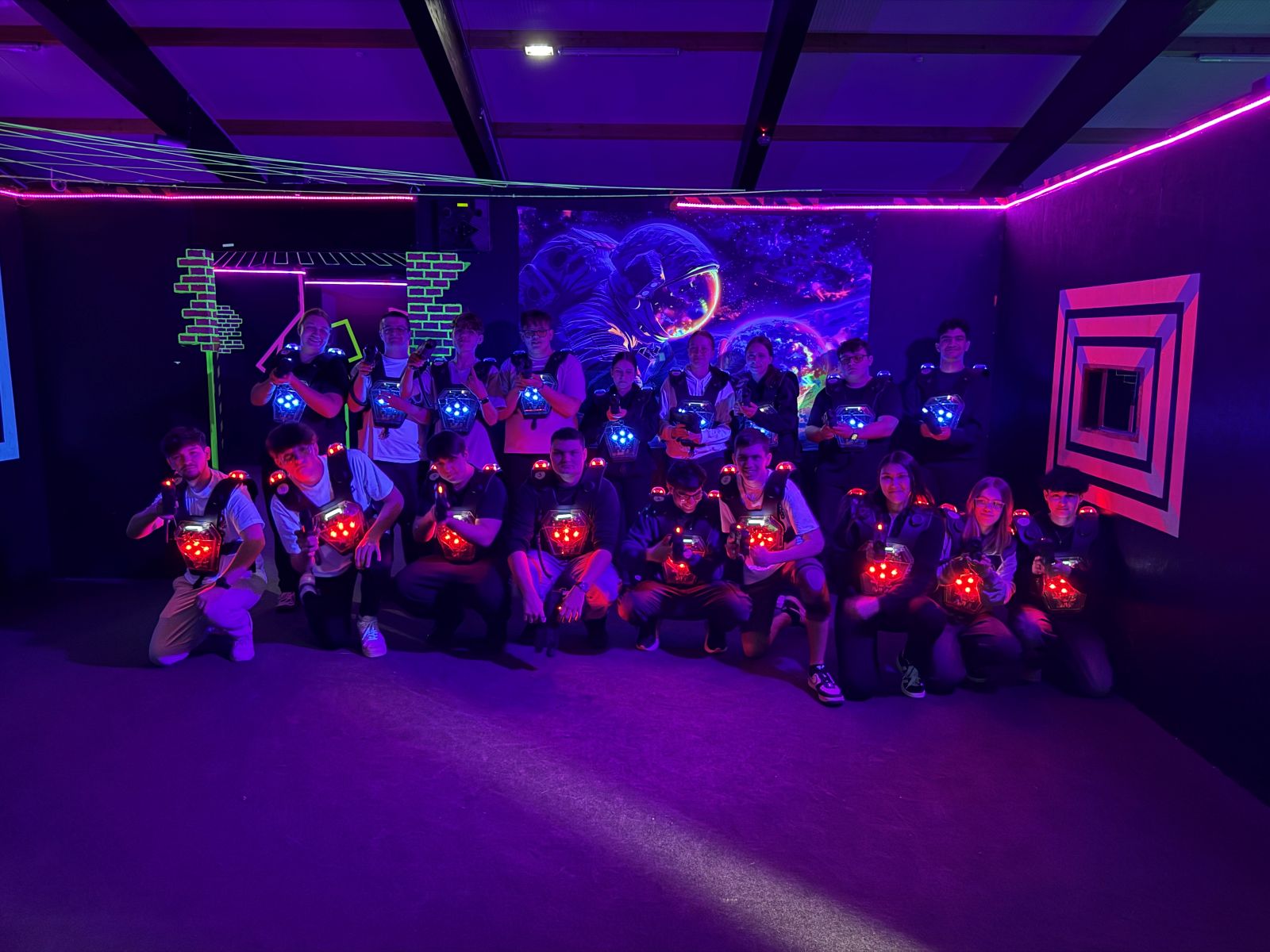 Eine große Gruppe von Schülern kniet und steht im dunklen Lasertag-Spielfeld, ihre Westen leuchten in Rot und Blau.