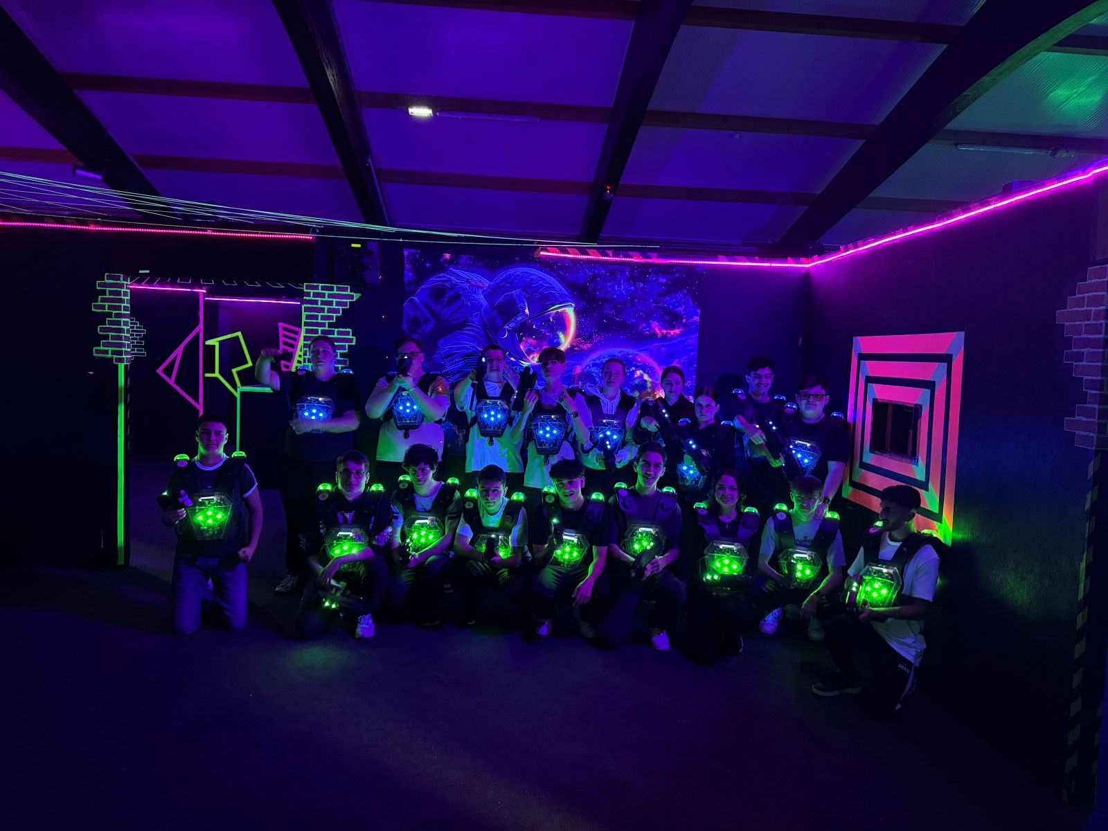 Schülergruppe posiert im dunklen Lasertag-Spielfeld, ihre Westen leuchten in Grün und Blau, umgeben von neonfarbenen Wandmustern.
