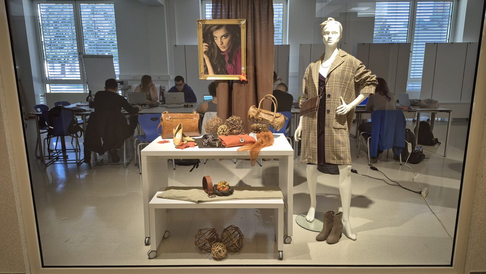Das Schaufenster widmet sich dem Thema Handtaschen. Eine Puppe, die einen karierten Mantel trägt, präsentiert eine elegante Handtasche. Mehrere andere Handtaschen sind auf einem weißen Präsentations-Tisch zusammen mit Tüchern, orangefarbenen Blättern und dekorativen Kugeln aus Naturmaterialien arrangiert.