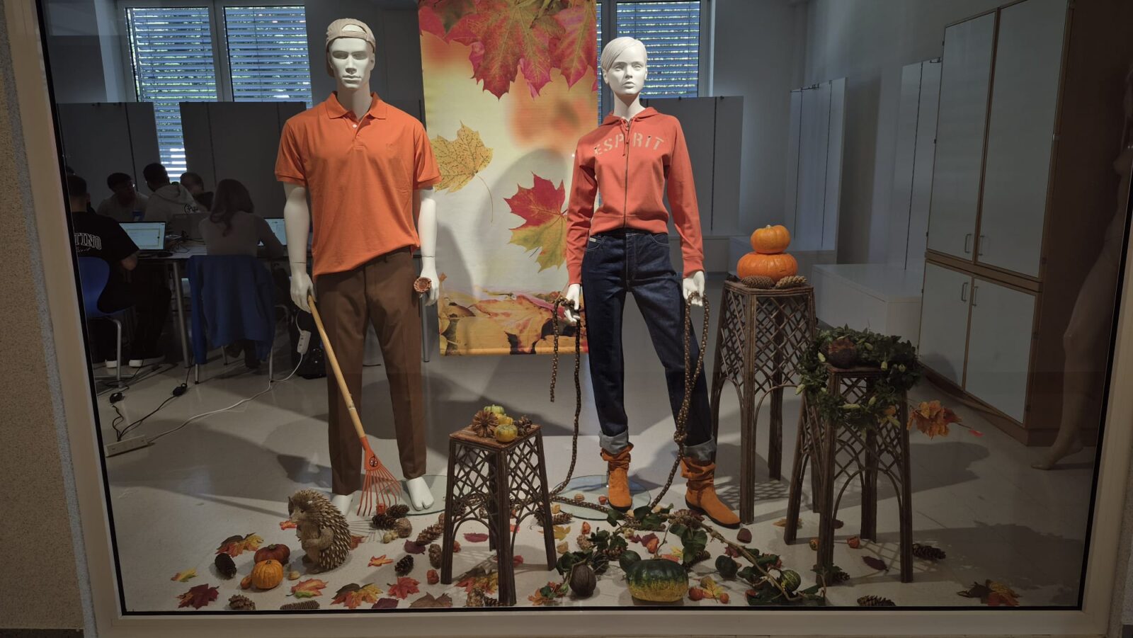 Das chaufenster steht ganz im Zeichen der Gartenarbeit im Herbst. Zwei Mannequins sind als Gärtner:innen gekleidet, mit einem Rechen und herbstlichem Laubwerk, das auf dem Boden verstreut ist. Das Schaufenster ist mit Kürbissen, Blättern und einem kleinen Igel verziert, was eine fröhliche und einladende Atmosphäre schafft.
