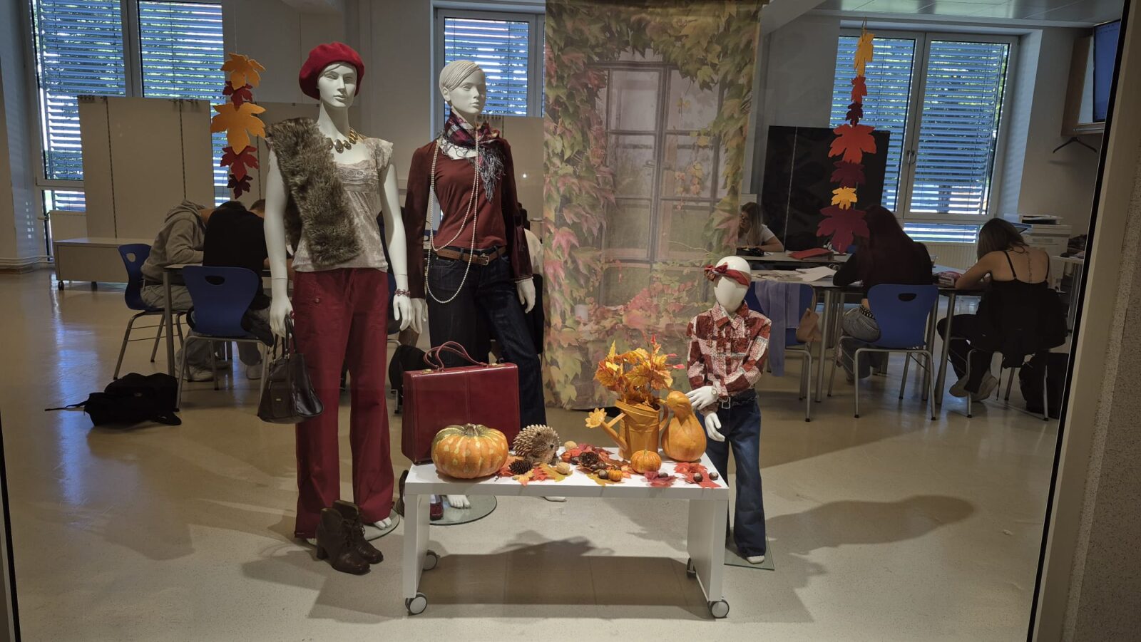 Ein Schaufenster entführt uns in die Welt der Herbstmode mit Kürbissen. Zwei Mannequins präsentieren stilvolle Herbst-Outfits, geschmückt mit typischen Herbstblättern. Neben einem Kürbis-Arrangement, einem kleinen Igel und einem Vogelhäuschen steht auch eine kleine Kinderfigur, die lässig in Jeans und kariertem Hemd gekleidet ist.