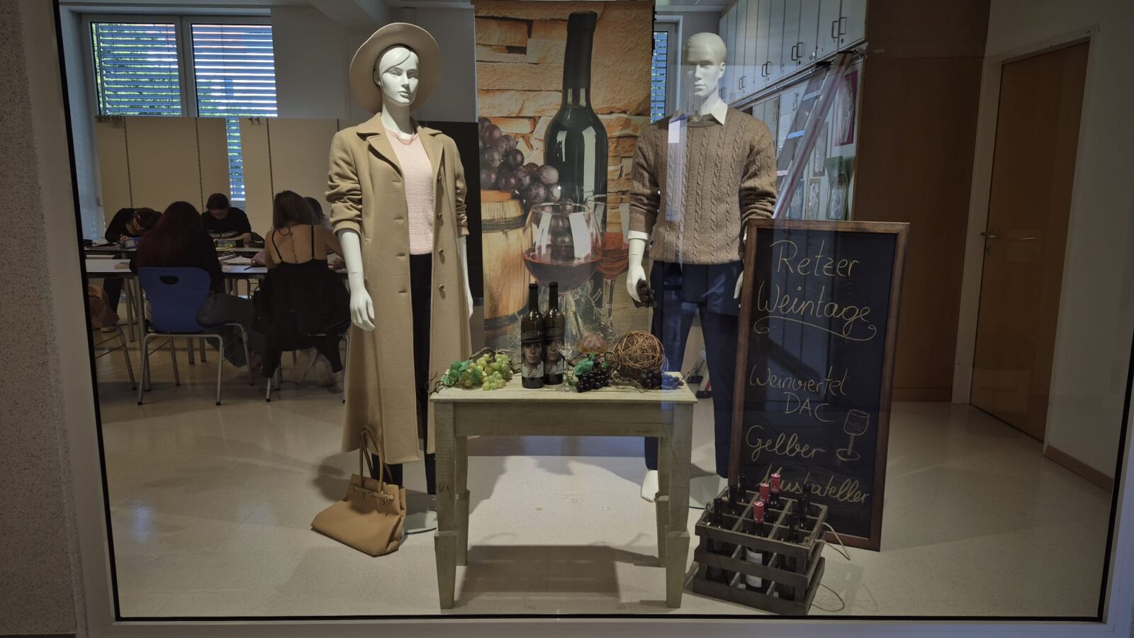 In diesem Schaufenster reht sich alles um das Thema Mode und Wein. Eine weibliche Figur trägt einen eleganten, hellen Mantel mit Hut, während ihr männliches Pendant einen beigen Pullover mit Zopfmuster trägt. Passend zum Thema schmückt eine Weinflasche mit Trauben und einem Weinglas das Schaufenster. Ein rustikales Kreidetafel-Schild weist auf die „Retzer Weintage“ hin, was dem Ganzen eine authentische Note verleiht.