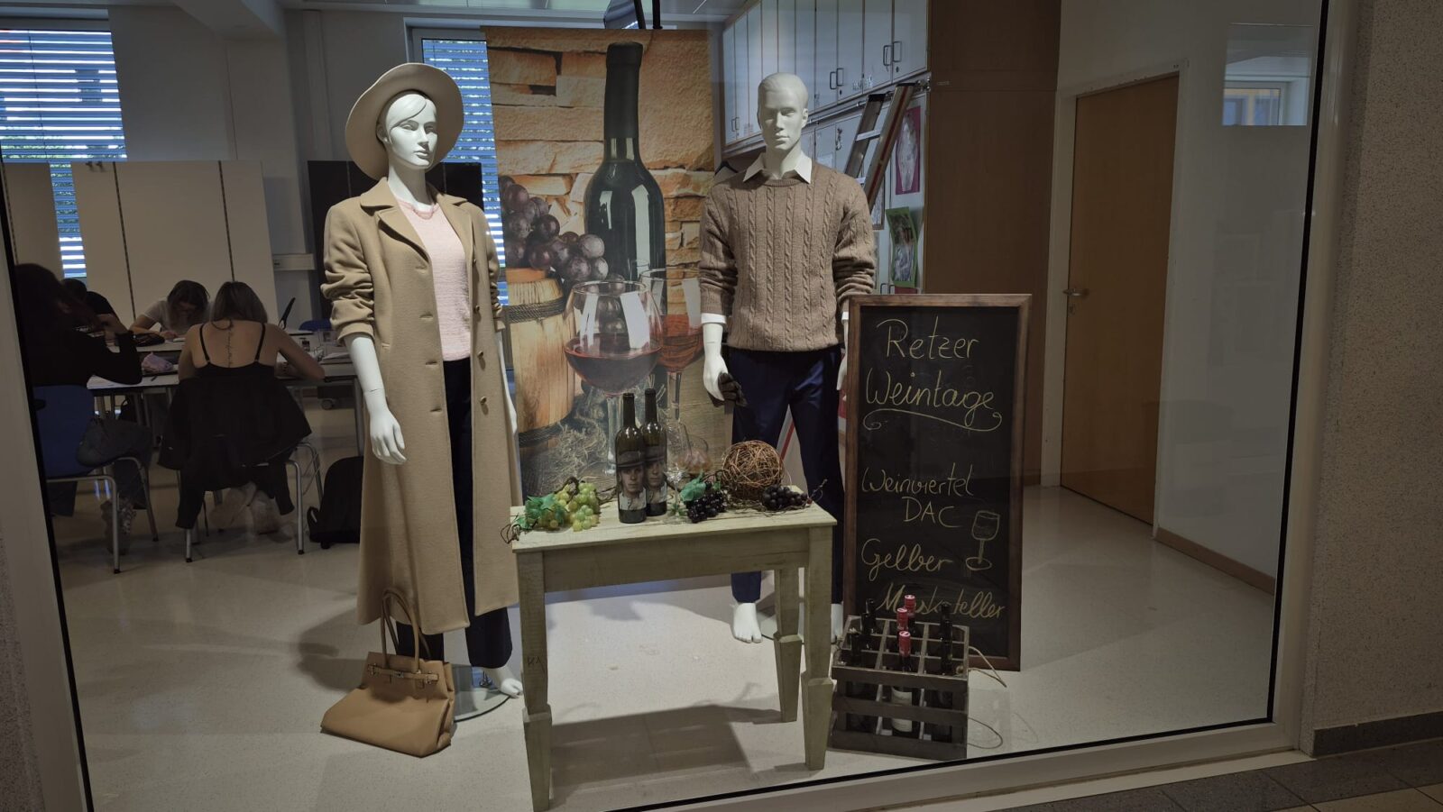 reht sich alles um das Thema Mode und Wein. Eine weibliche Figur trägt einen eleganten, hellen Mantel mit Hut, während ihr männliches Pendant einen beigen Pullover mit Zopfmuster trägt. Passend zum Thema schmückt eine Weinflasche mit Trauben und einem Weinglas das Schaufenster. Ein rustikales Kreidetafel-Schild weist auf die „Retzer Weintage“ hin, was dem Ganzen eine authentische Note verleiht.
