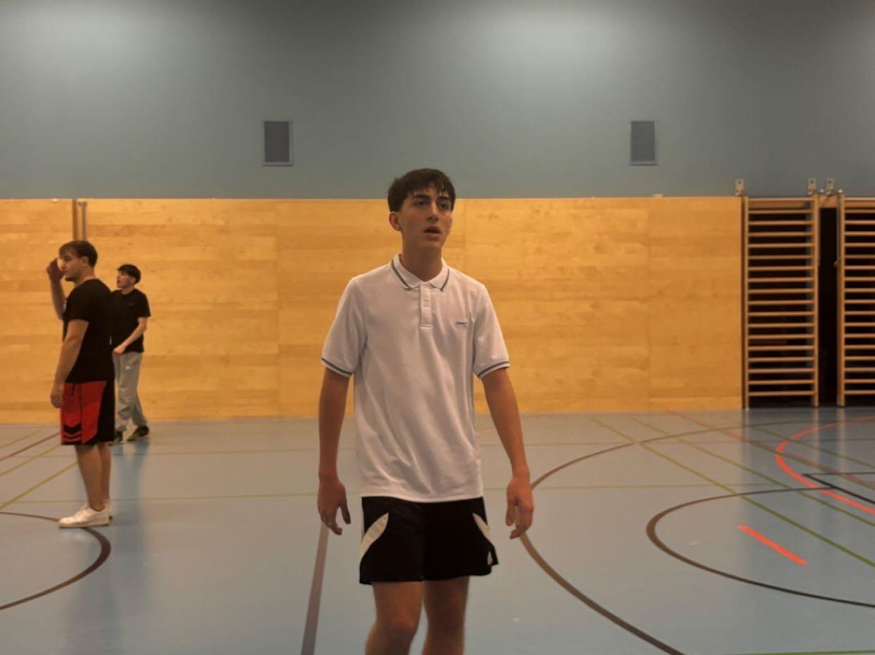 Spieler beim Match Gerne, hier ist der Alternativtext für das neueste Bild in Ihrem Stapel, das noch keinen Alternativtext erhalten hat: image_37b9e2.jpg. Alternativtext für image_37b9e2.jpg (Junge im Turnsaal): Ein junger Mann in einem weißen Polo-Shirt und schwarzen Shorts steht auf einem blauen Sportboden in einer Turnhalle. Im Hintergrund sind weitere Personen und Turngeräte zu sehen.