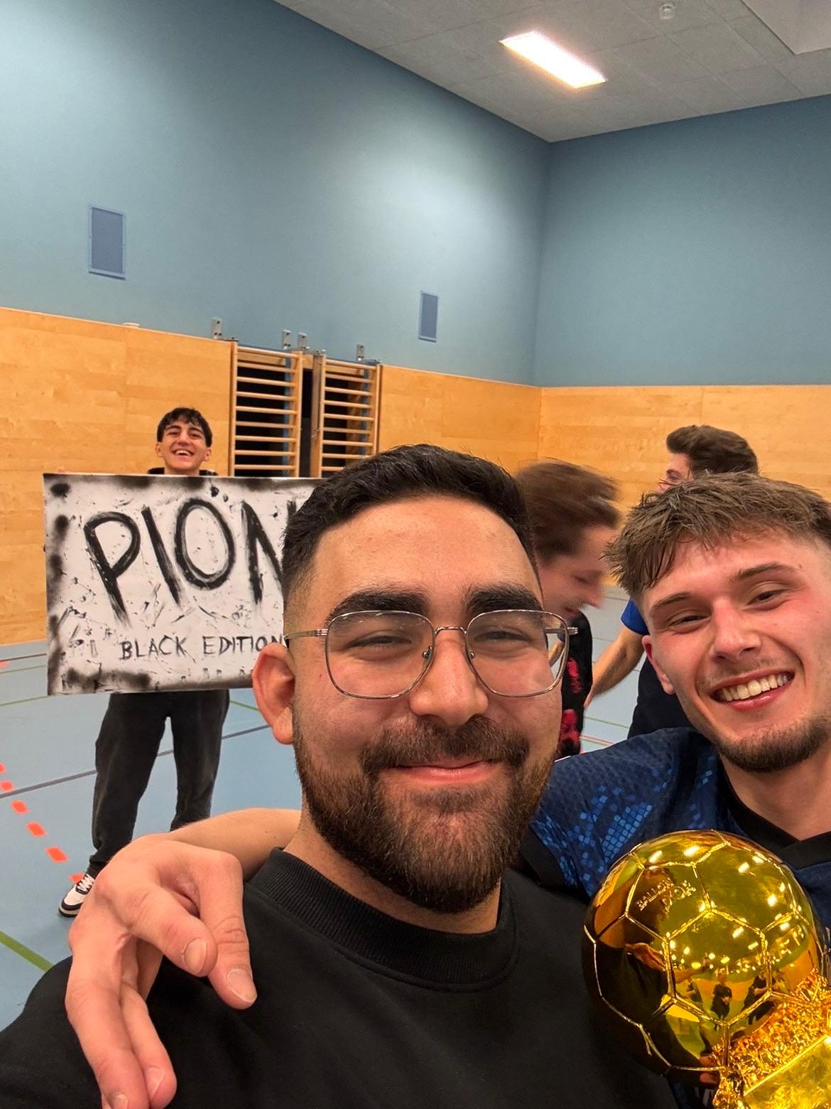 Freude nach dem Turnier Ein Selfie von zwei jungen Männern, die in die Kamera lächeln. Einer hält eine goldene Trophäe, der andere trägt eine Brille und hält im Hintergrund ein Schild mit der Aufschrift "PION BLACK EDITION" hoch, während weitere Personen im Turnsaal verschwommen sind.