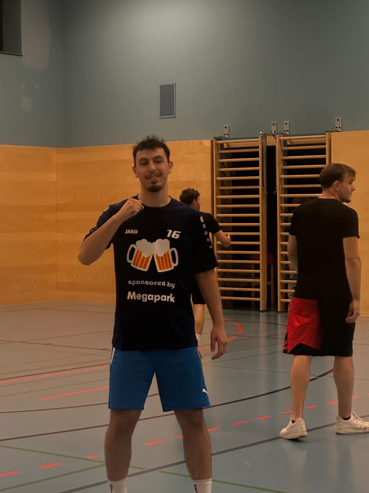 In der Turnhalle Ein junger Mann im Turnsaal posiert in einem dunklen Trikot mit der Aufschrift "Sponsored by Megapark" und blauen Sporthosen. Im Hintergrund sind weitere Personen und Turngeräte zu sehen.