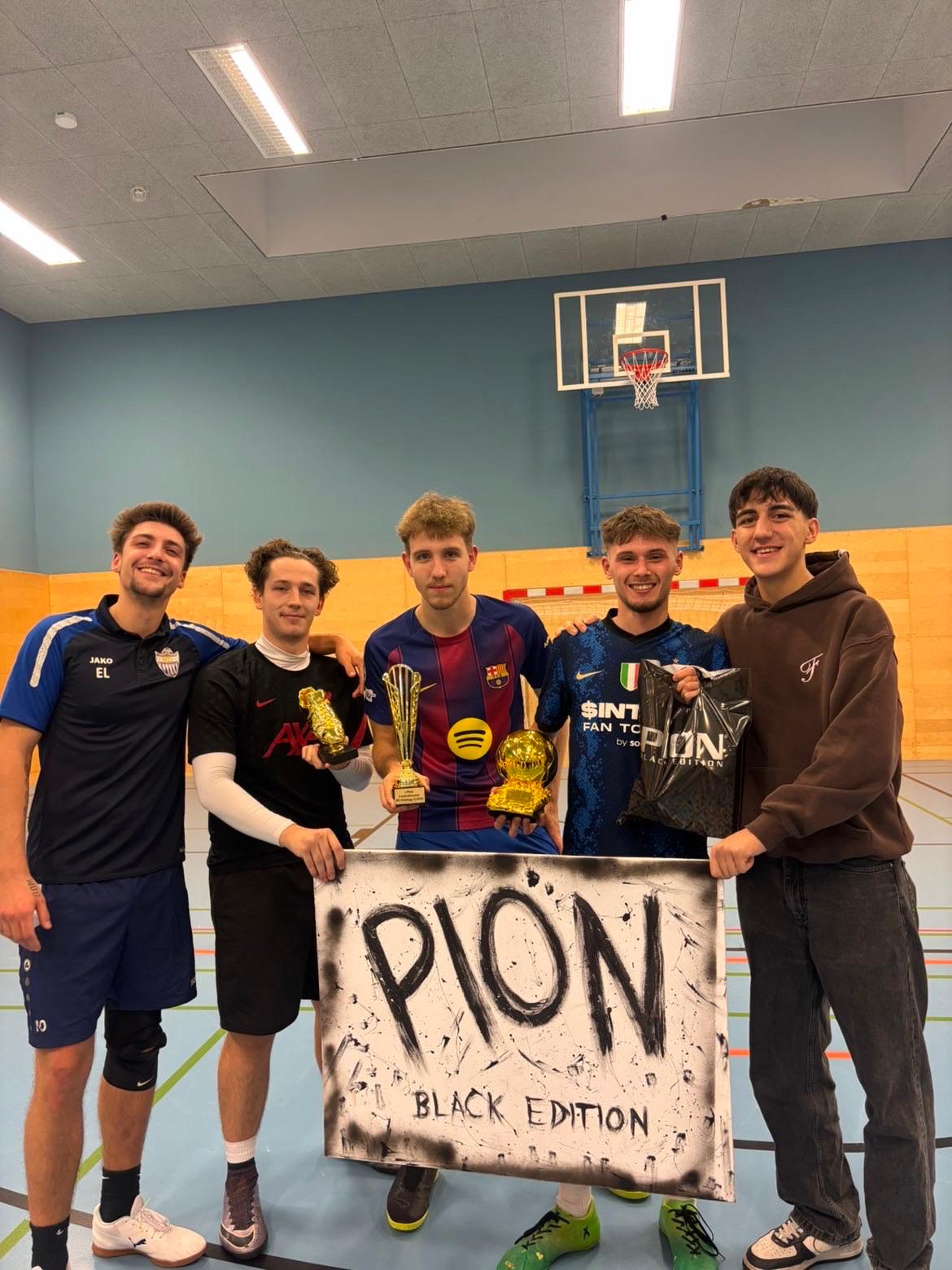 Team und Sponsor Fünf junge Männer, einige in Fußballtrikots und Sportkleidung, posieren in einer Turnhalle. Sie halten einen Siegerschild mit der Aufschrift "PION BLACK EDITION" und zwei goldene Trophäen in die Höhe.
