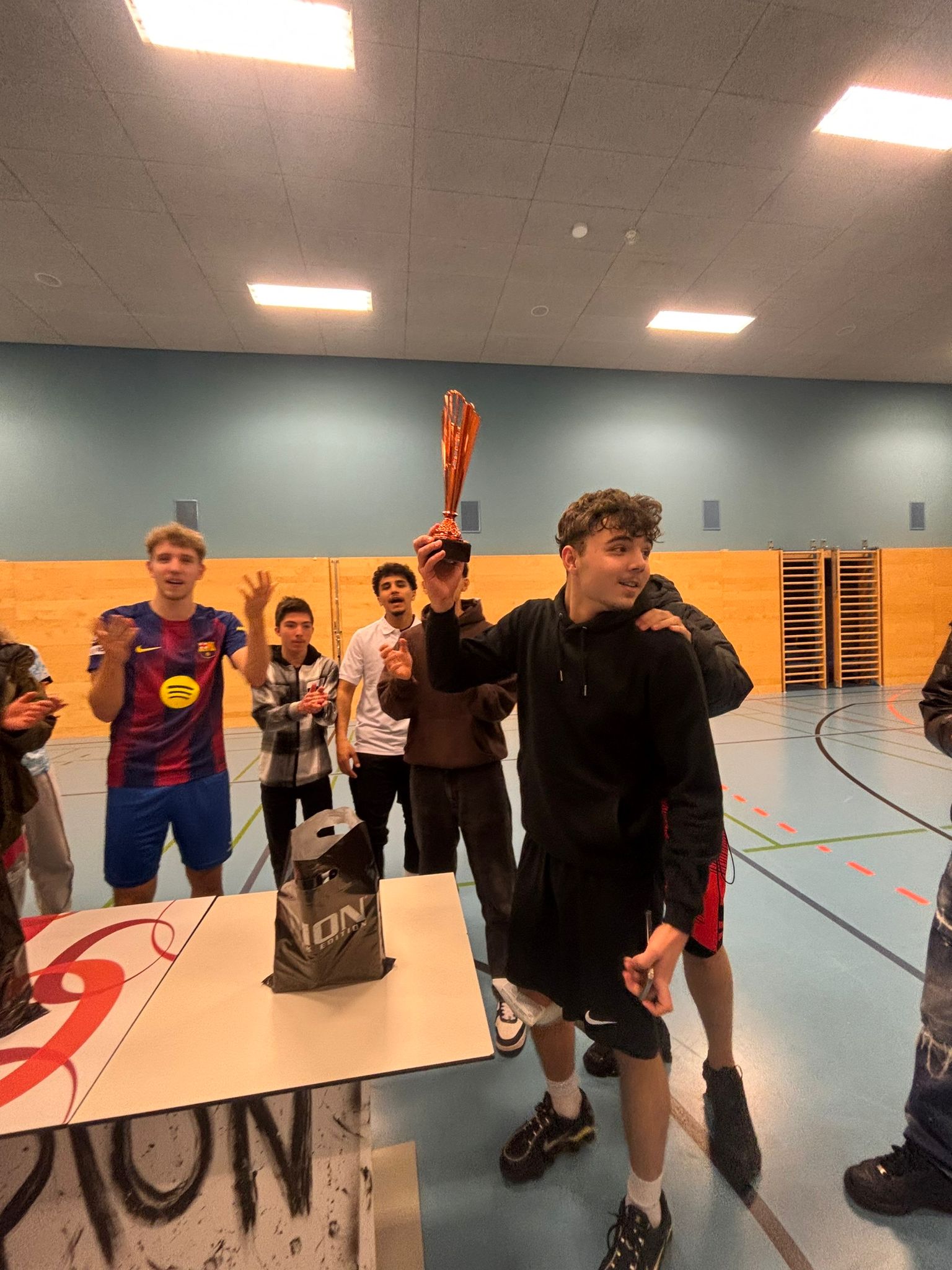 nach dem Turnier Ein junger Mann im Vordergrund hält eine Trophäe in die Höhe und jubelt, während eine Gruppe von jungen Männern im Turnsaal um einen kleinen Tisch herumsteht und applaudiert. Auf dem Tisch steht ein schwarzer Beutel mit dem "PION" Logo.