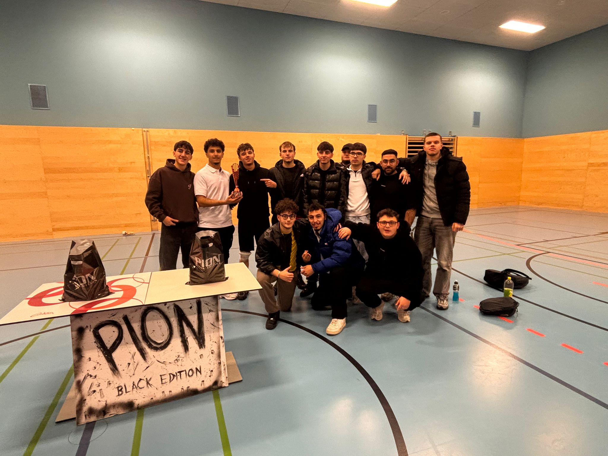 Das Team Eine Gruppe junger Männer posiert in einer Sporthalle um einen kleinen Tisch, auf dem das "PION BLACK EDITION" Logo zu sehen ist. Einige tragen schwarze Jacken und Pullover und lächeln in die Kamera.