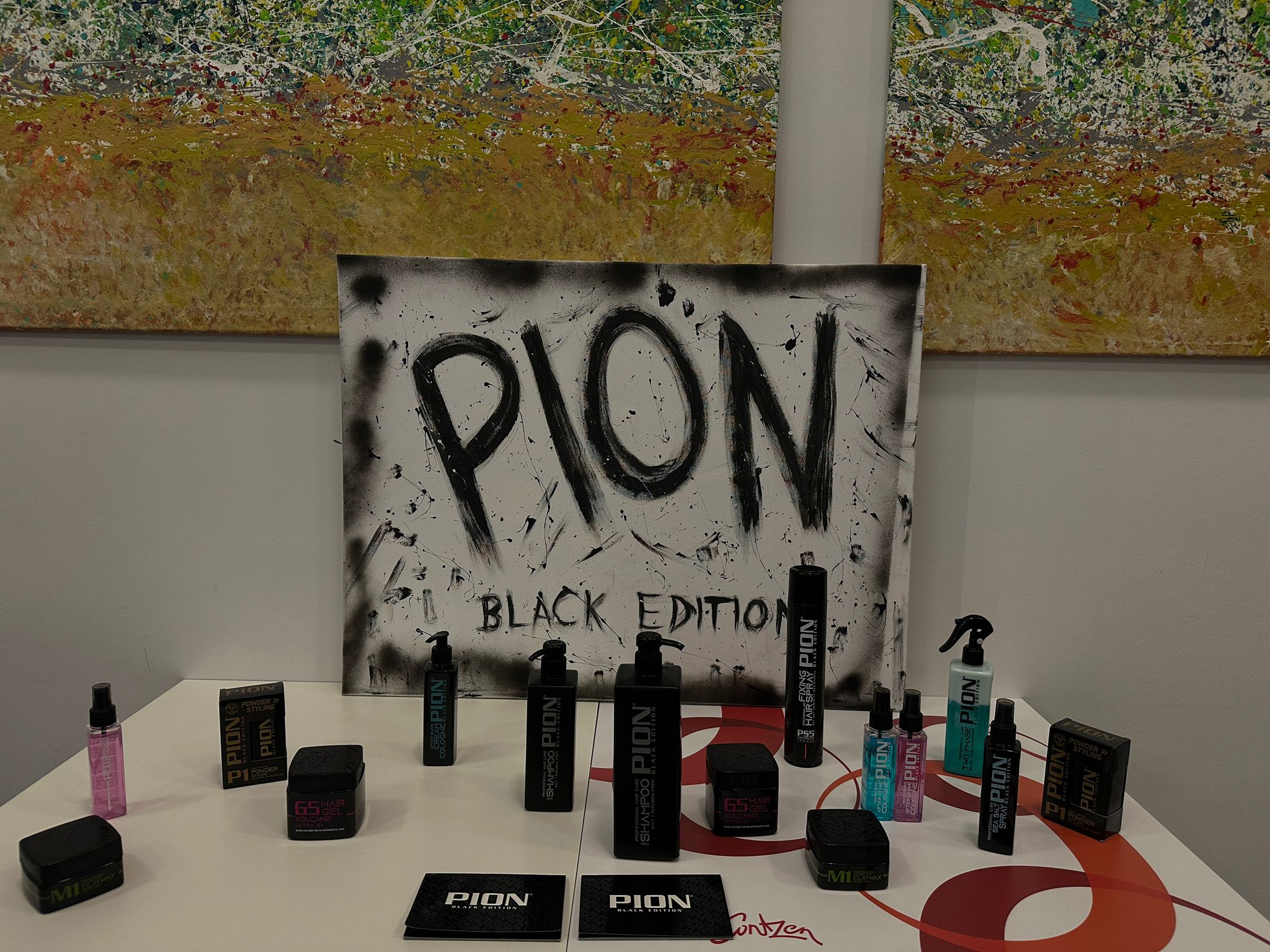 Unser Sponsor Eine Nahaufnahme eines Tisches, auf dem verschiedene schwarze und farbige Flaschen mit dem Logo "PION" ausgestellt sind. Dahinter steht eine weiße Leinwand mit dem handgemalten Schriftzug "PION BLACK EDITION" in Schwarz und ein abstraktes Gemälde hängt im Hintergrund.