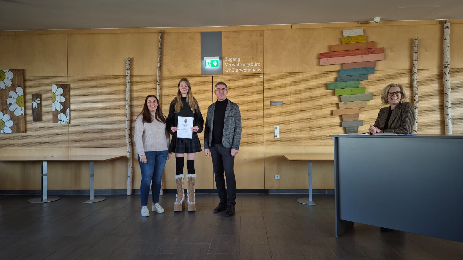 3BM1 - KV jar Zwei Erwachsene und eine junge Frau, die eine Urkunde hält, posieren lächelnd in einem Flur mit Holzverkleidung für ein Foto. Eine weitere erwachsene Frau sitzt an einem Tisch auf der rechten Seite.