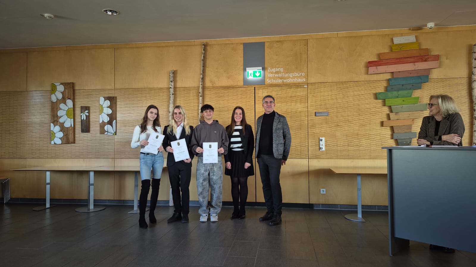 3GH1 - KV bka (am Bild: skm) Vier junge Erwachsene, die Urkunden halten, posieren mit einem erwachsenen Mann in einem Flur mit Holzverkleidung für ein Foto. Eine weitere erwachsene Frau sitzt an einem Tisch auf der rechten Seite.