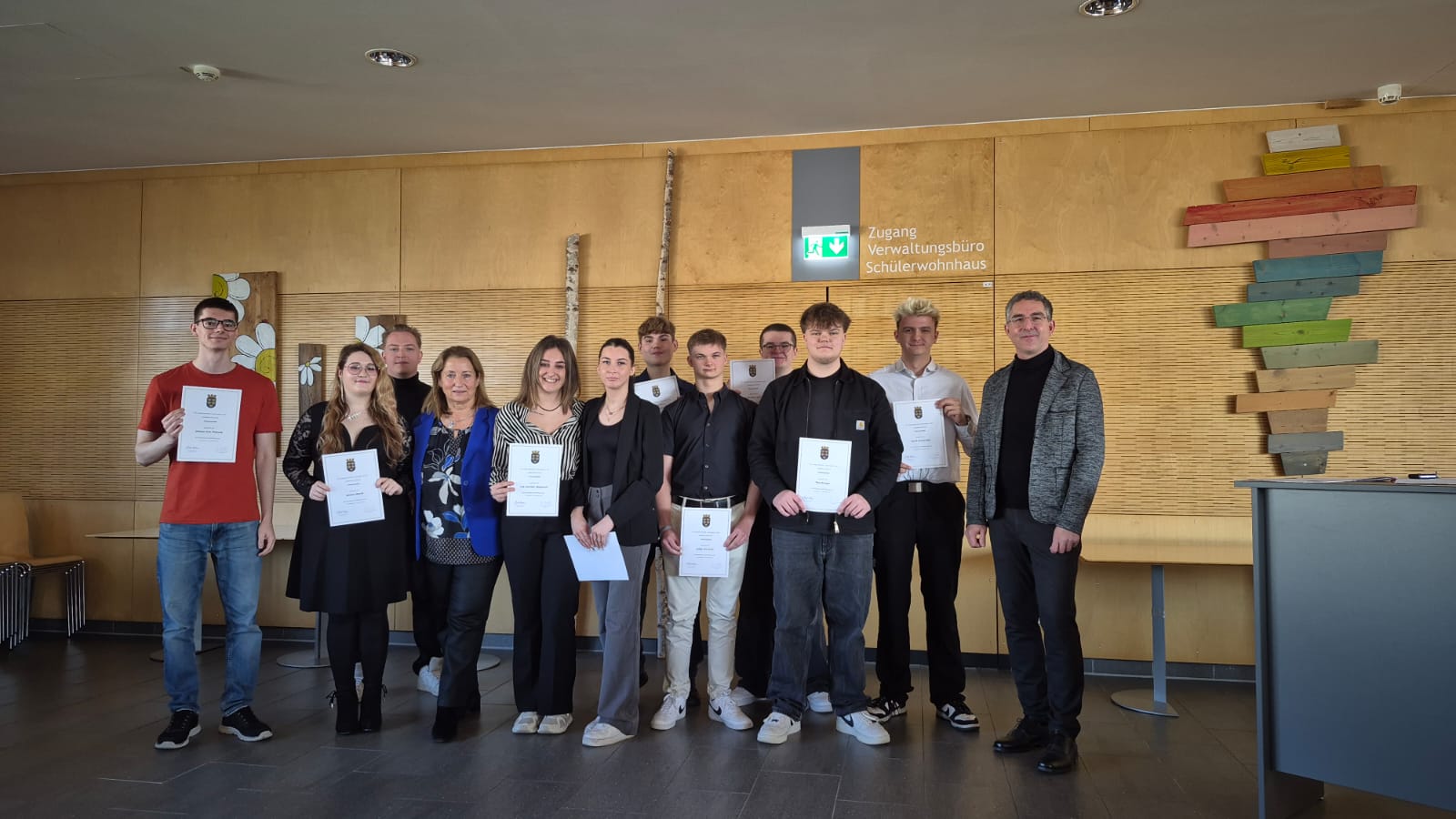3EL - KV scr Eine Gruppe von acht jungen Erwachsenen, die Urkunden halten, posiert mit einem erwachsenen Mann in einem Flur mit Holzverkleidung für ein Foto. Eine weitere erwachsene Frau sitzt an einem Tisch auf der rechten Seite.