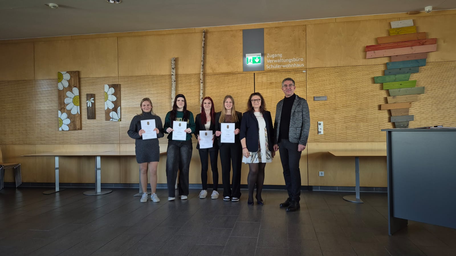 3T1 - KV neb Fünf junge Frauen, die Urkunden halten, posieren mit einem erwachsenen Mann in einem Flur mit Holzverkleidung für ein Foto. Eine weitere erwachsene Frau sitzt an einem Tisch auf der rechten Seite.
