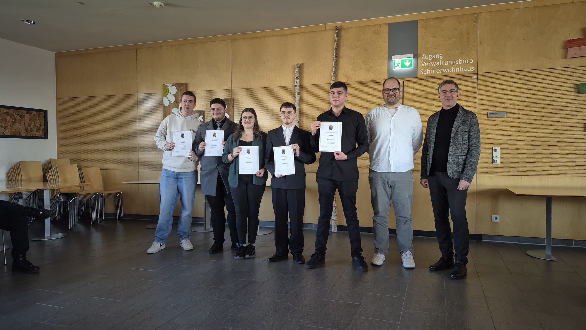 3LH1 - KV mus Fünf junge Erwachsene, die Urkunden halten, posieren mit zwei erwachsenen Männern in einem Flur mit Holzverkleidung für ein Foto.