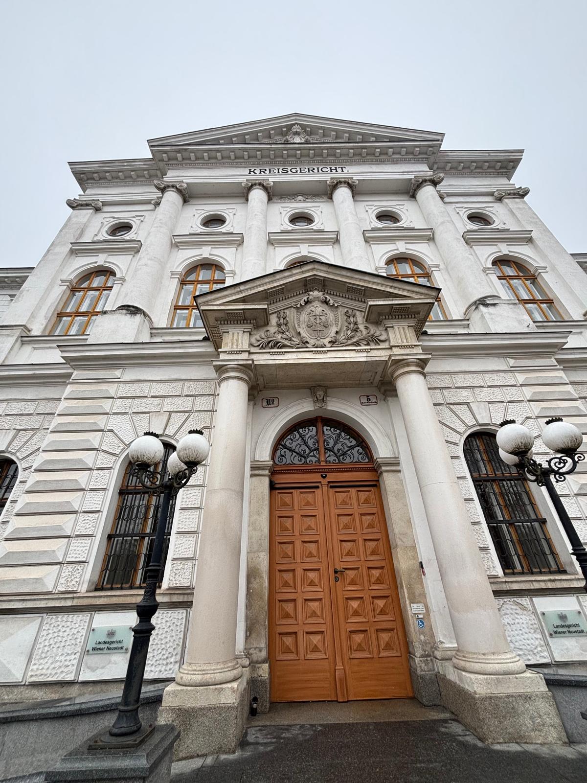 Landesgericht Wiener Neustadt Die Außenansicht eines großen, historischen Steingebäudes bei Tageslicht. Die Fassade ist hellgrau und verfügt über zahlreiche regelmäßig angeordnete Fenster mit dunklen Rahmen. Ein zentraler Gebäudeteil ragt leicht hervor und ist mit einem flachen Dach sowie einer schlichten Attika abgeschlossen. Vor dem Gebäude befindet sich ein gepflasterter Bereich und ein Gehweg. Die Architektur wirkt sachlich und monumental.