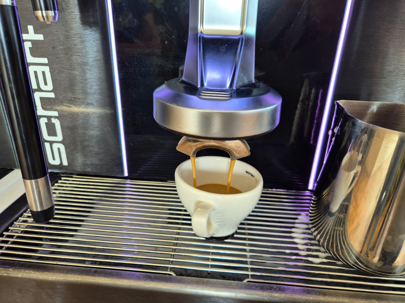Exkursion_Schärf 10 Eine Espressomaschine der Marke Schärf bereitet gerade einen Kaffee zu. Aus dem Auslauf fließt frisch gebrühter Espresso in eine weiße Tasse, daneben steht ein Edelstahl-Milchkännchen auf dem Gitterrost.