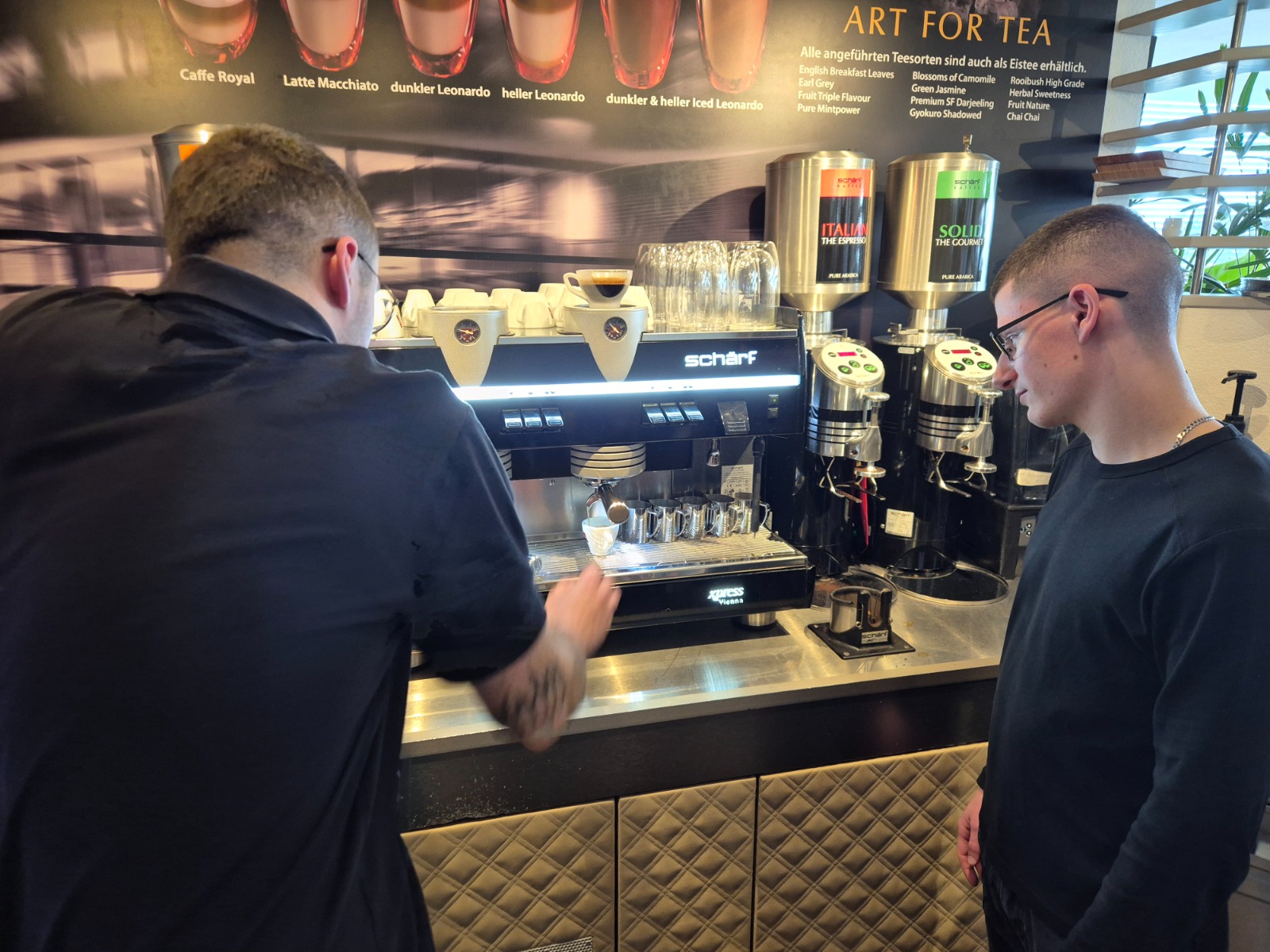 Exkursion_Schärf 11 Zwei Personen stehen vor einer Kaffeemaschine in einem Vorführraum. Eine Person zeigt der anderen, wie man Kaffee zubereitet. Im Hintergrund sind verschiedene Kaffeebezeichnungen und Teesorten zu sehen.