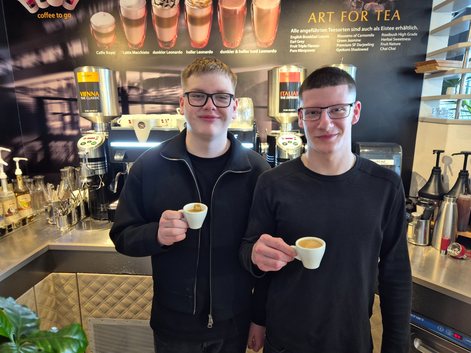 Exkursion_Schärf 12 Zwei Schüler:innen stehen in einer Kaffeebar und halten jeweils eine kleine Tasse Espresso in der Hand. Im Hintergrund sind Kaffeemaschinen und Getränkespender zu sehen.