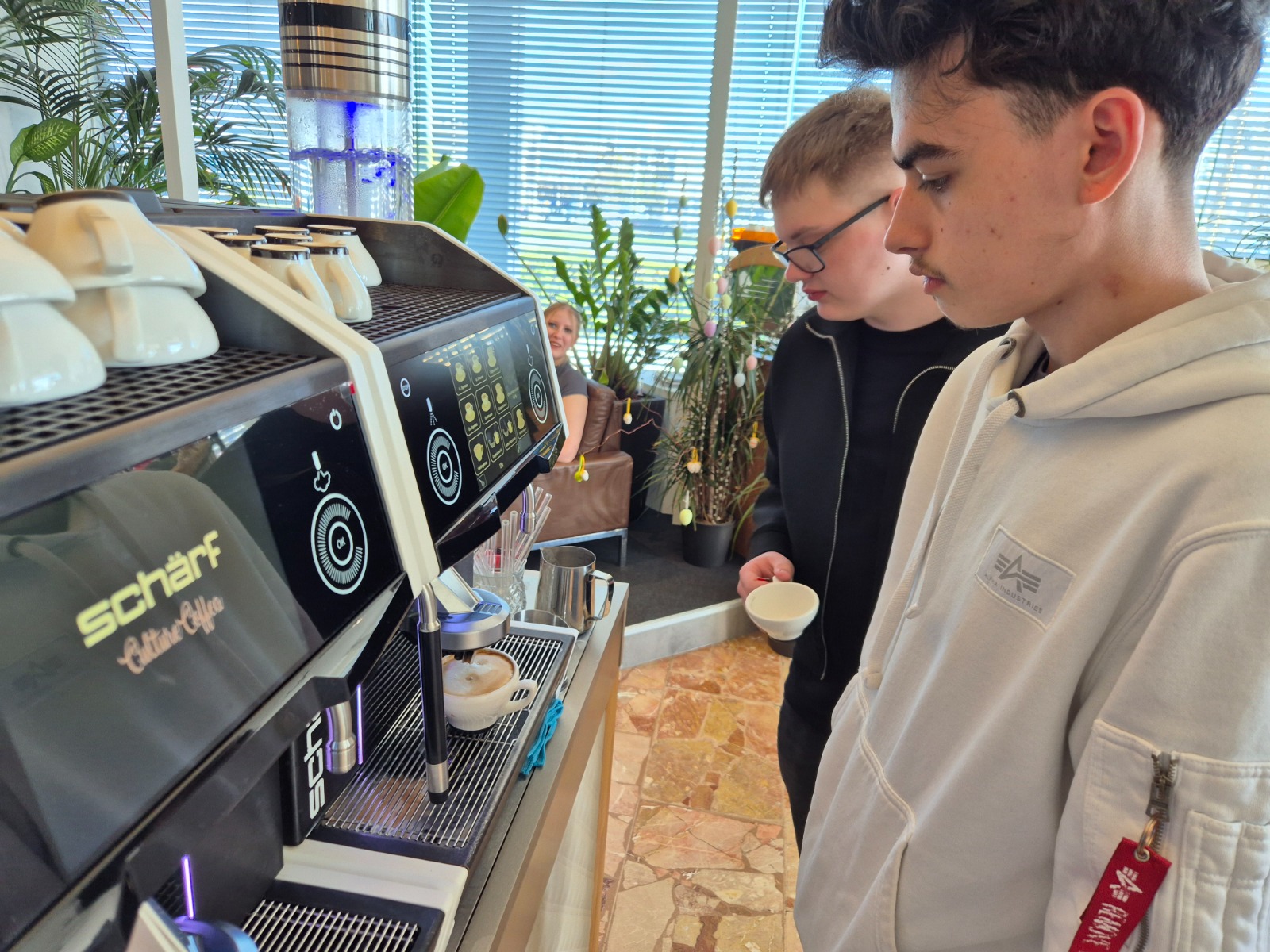 Exkursion_Schärf 13 Zwei Schüler:innen stehen an einer modernen Kaffeemaschine und beobachten aufmerksam die Zubereitung eines Cappuccinos während einer Exkursion zur Firma Schärf.