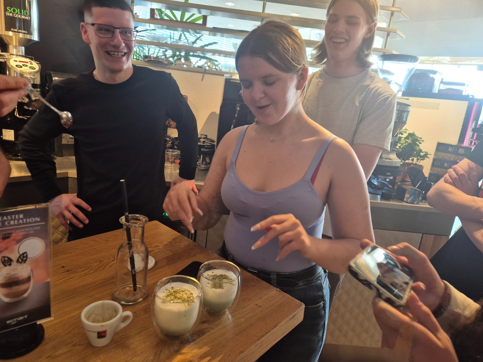 Exkursion_Schärf 15 Eine Gruppe von Schüler:innen steht in einem Café an einer Theke. Eine Schülerin zeigt gerade zwei dekorierte Matcha Latte, während andere zusehen und fotografieren. Die Szene wirkt lebendig und zeigt praktischen Unterricht im Rahmen einer Exkursion.