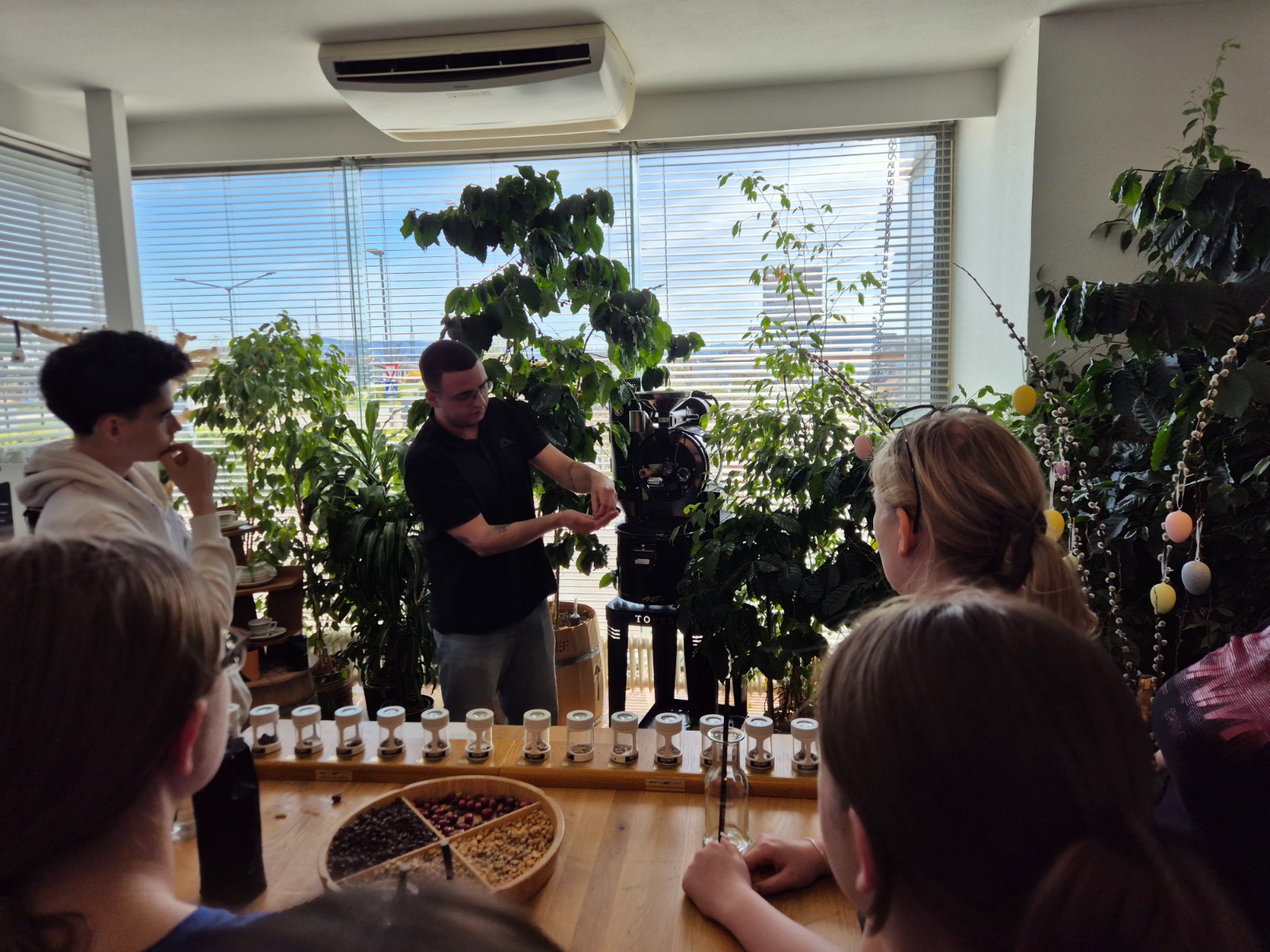 Exkursion_Schärf 3 Eine Gruppe von Schüler:innen verfolgt eine Vorführung zum Thema Kaffee in einem hellen Raum mit vielen Pflanzen.