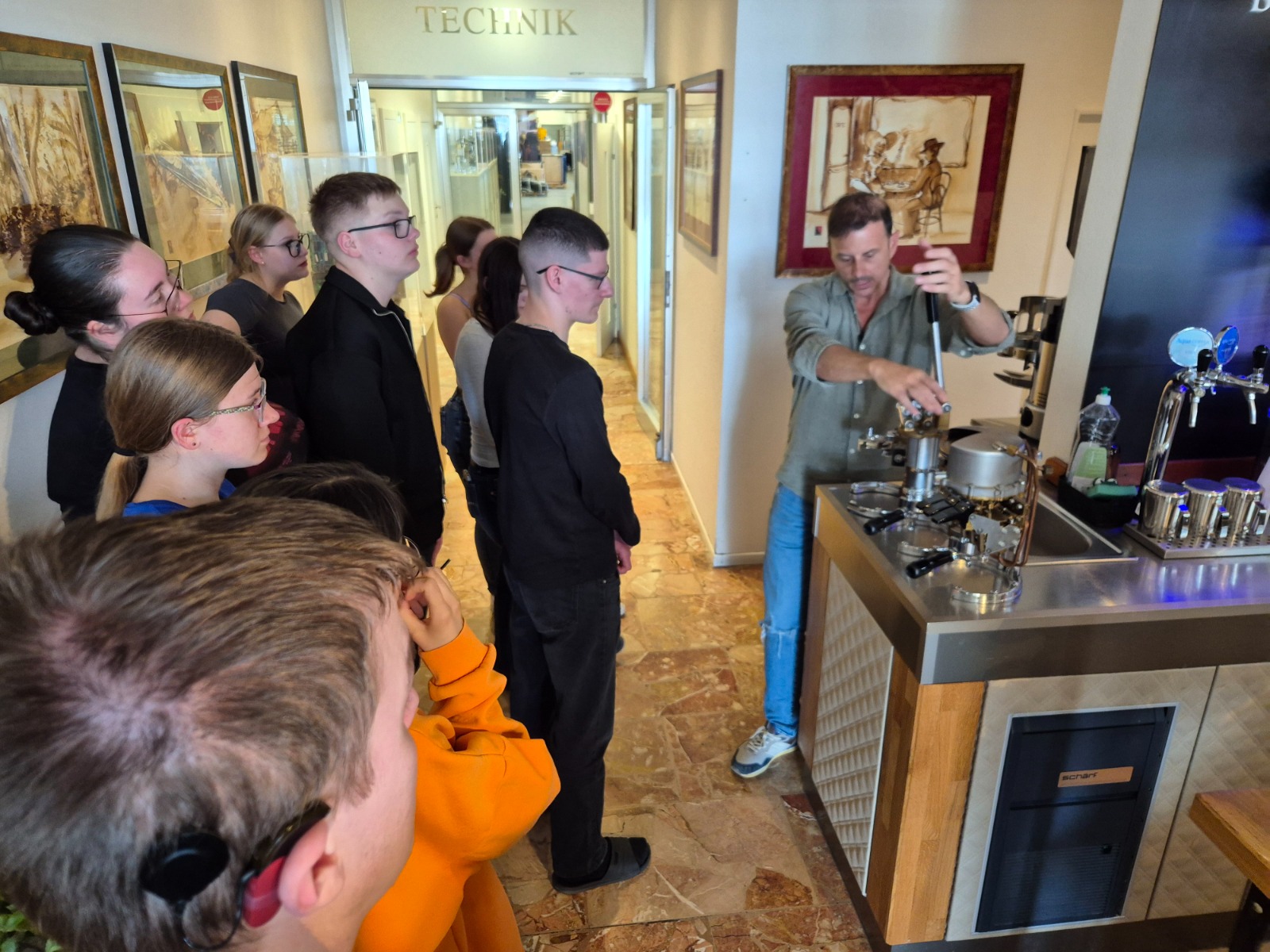 Exkursion_Schärf 7 Eine Gruppe von Schüler:innen steht um einen Mitarbeiter, der an einer Kaffeemaschine zeigt, wie die Bedienung funktioniert. Die Jugendlichen beobachten aufmerksam die Demonstration.