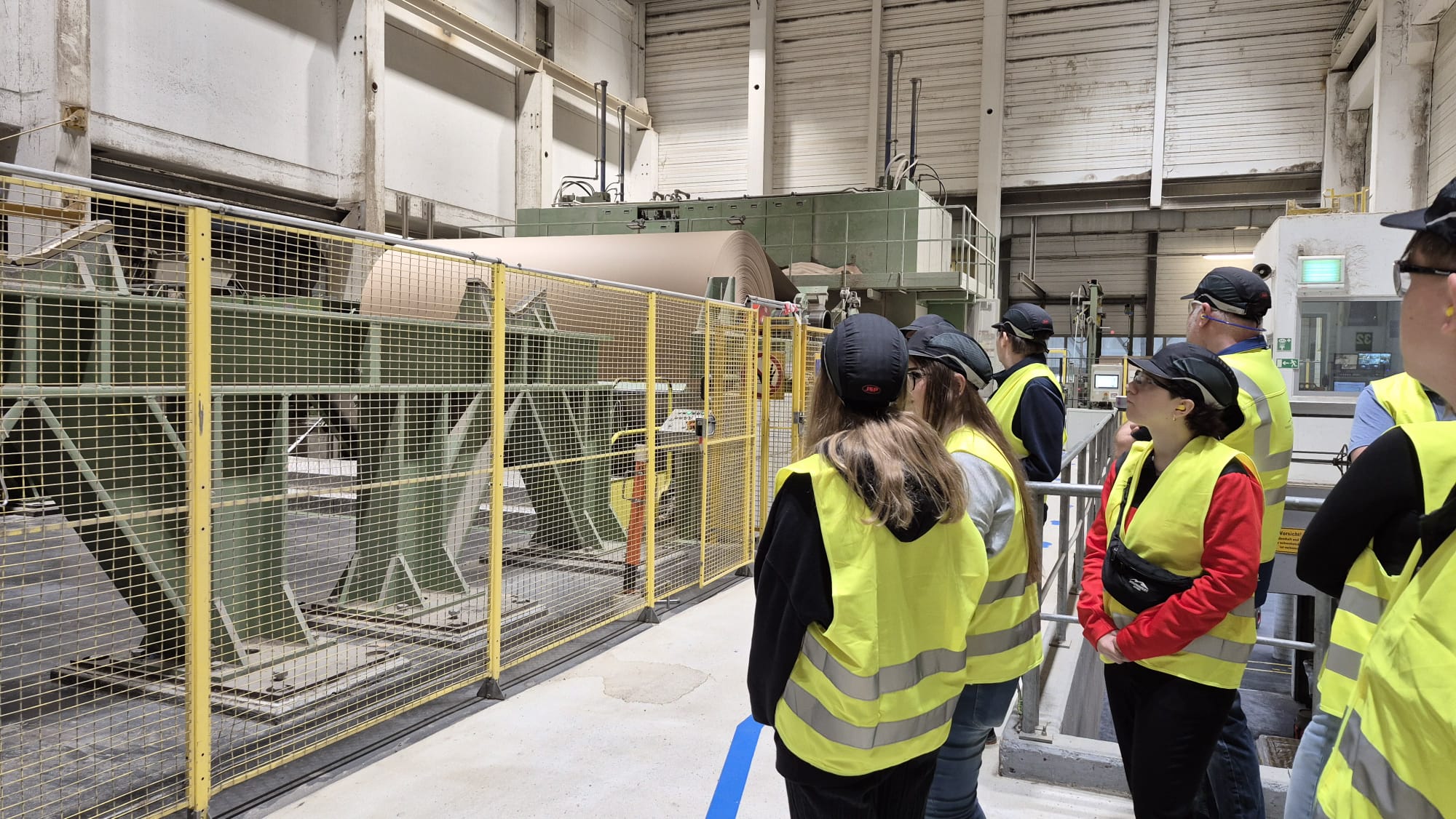 Papierfabrik Hamburger Containerboard GmbH Pitten Eine Gruppe von Personen mit Sicherheitshelmen und neongelben Warnwesten steht in einer großen Industriehalle vor einer Papiermaschine. Die Maschine ist teilweise durch ein gelbes Metallgitter von den Personen getrennt. In der Maschine ist eine große, braune Papierrolle sichtbar. Die Umgebung ist von industriellen Strukturen und hohen Wänden geprägt..