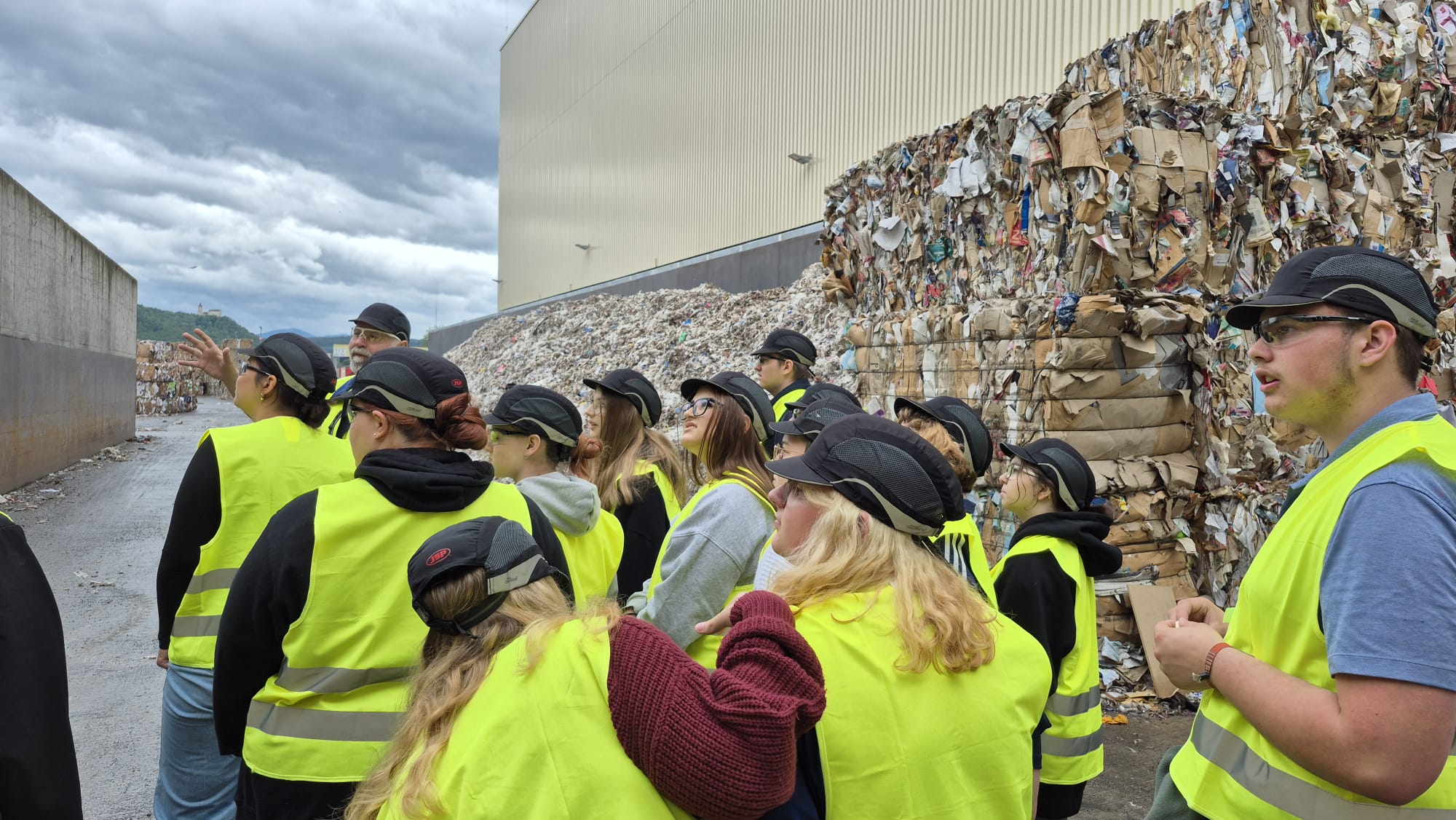 Papierfabrik Hamburger Containerboard GmbH Pitten Eine Gruppe von Personen mit Sicherheitshelmen und neongelben Warnwesten steht auf einem Fabrikgelände. Eine Person mit erhobenem Arm zeigt auf einen Bereich außerhalb des Bildfeldes, während die Gruppe in diese Richtung blickt. Rechts im Bild sind hohe Stapel von gepressten, gebrauchten Kartonballen vor einem großen Fabrikgebäude aufgeschichtet. Der Boden ist grau, der Himmel bewölkt.