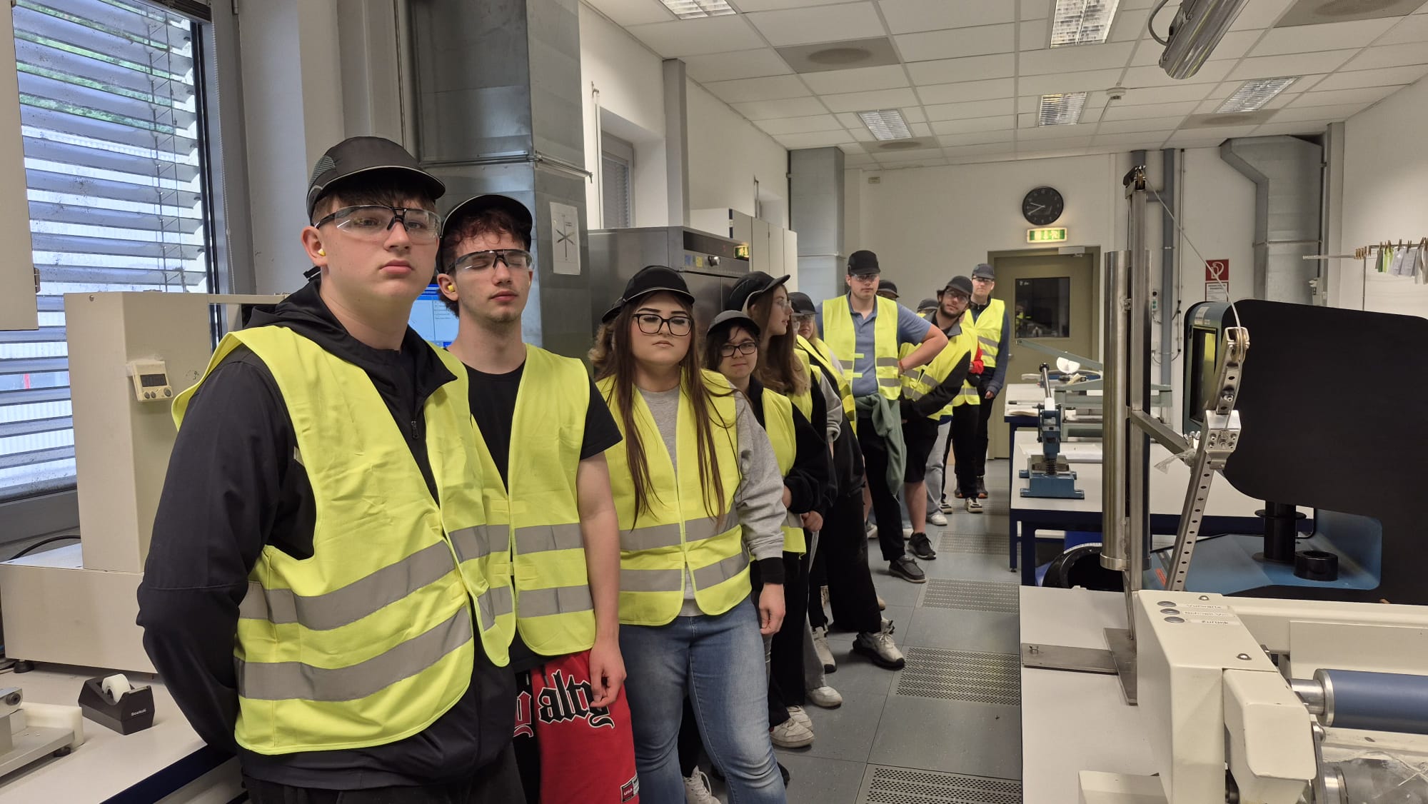 Papierfabrik Hamburger Containerboard GmbH Pitten Eine Gruppe von Personen mit Sicherheitshelmen und neongelben Warnwesten steht in einer Reihe in einem hellen Labor- oder Werkstattraum. Die Personen blicken mehrheitlich nach vorne. Der Raum ist mit verschiedenen technischen Geräten und Arbeitstischen ausgestattet. Im Hintergrund sind eine Tür, eine Wanduhr sowie ein Fenster mit Lamellenjalousien zu sehen.