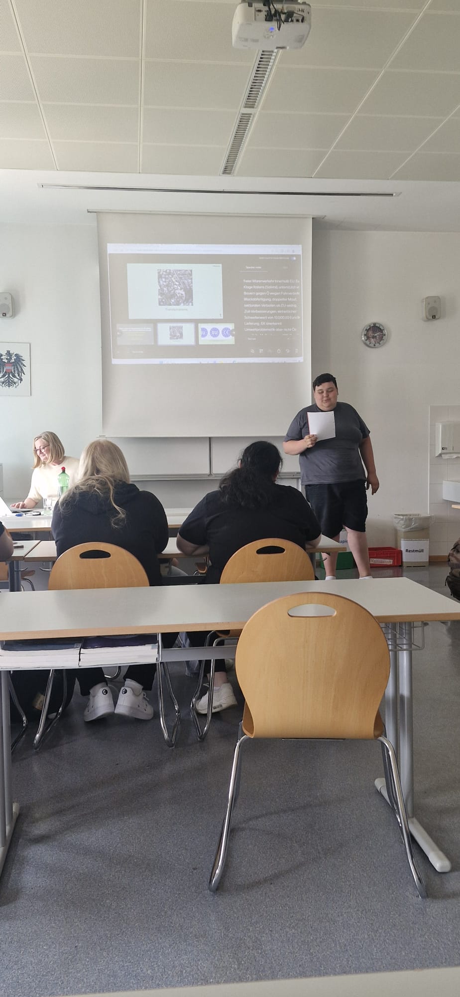 EU-Projekt_Teil2_1 Im Unterrichtsraum präsentieren Schülerinnen und Schüler vor der Klasse eine Arbeit. Auf der Leinwand ist eine Präsentation zu sehen, während eine Person vorne steht und einen Zettel in der Hand hält.