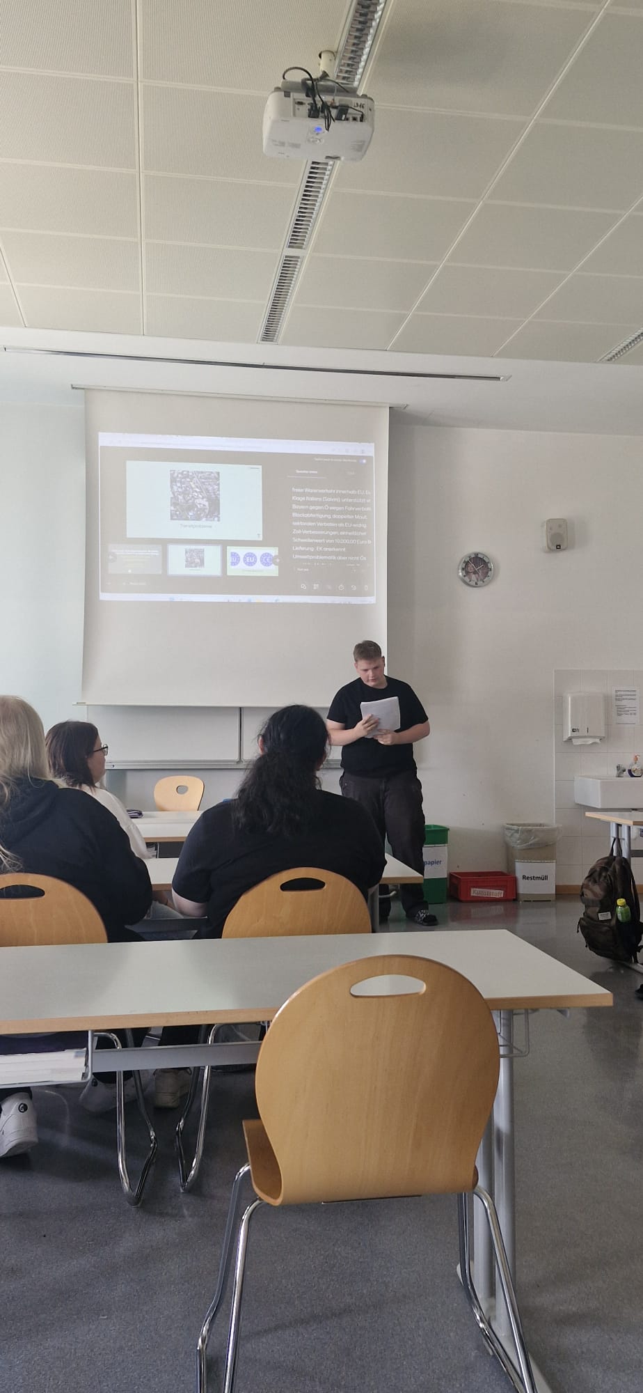EU-Projekt_Teil2_3 Im Unterrichtsraum präsentieren Schülerinnen und Schüler vor der Klasse eine Arbeit. Auf der Leinwand ist eine Präsentation zu sehen, während eine Person vorne steht und einen Zettel in der Hand hält.