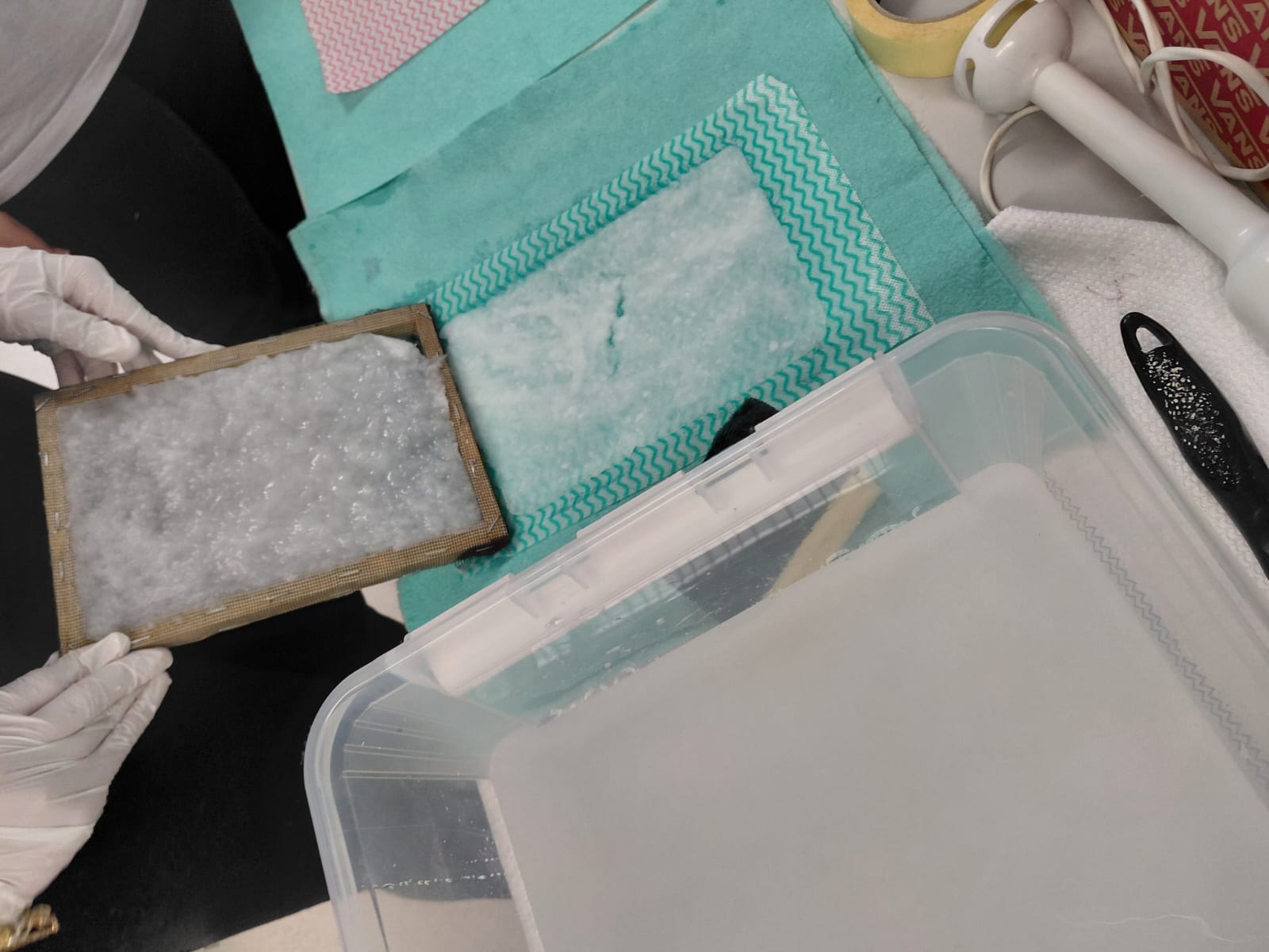 Workshop "Papierschöpfen" Zwei Hände mit Handschuhen halten einen Rahmen mit weißer Papiermasse an eine Arbeitsfläche, auf der bereits ein abgegautschtes Stück Papier liegt. Davor ein Behälter mit Pulpe.