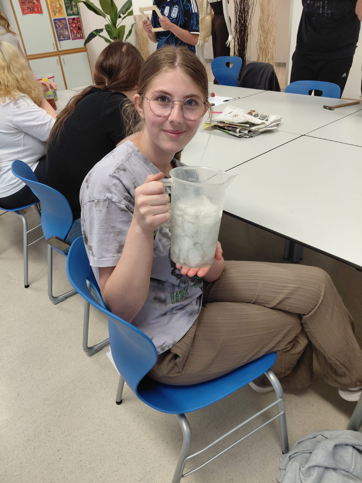 Schüler:innen der 1.PA beim Workshop "Papierschöpfen" Sitzende Person hält einen Plastikkrug mit Pulpe in der Hand und präsentiert ihn in einem Klassenraum.