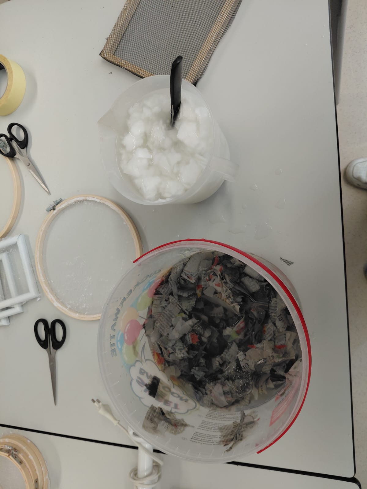 Workshop "Papierschöpfen" Eine Arbeitsfläche mit einem Kübel dunklen Papierbreis, einem Krug mit weißem Papierbrei, mehrere Rahmen und zwei Scheren.