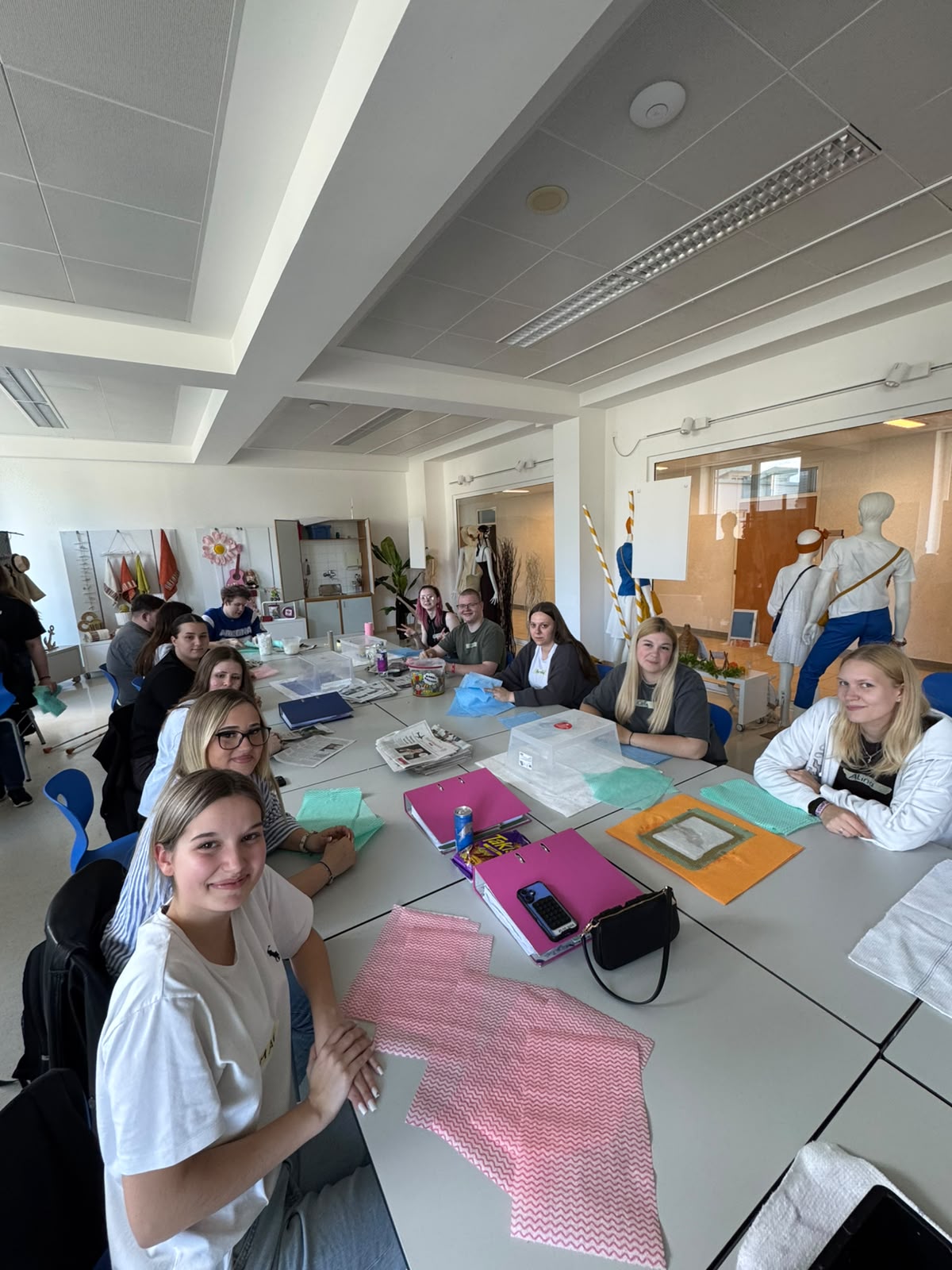 Die 3.PA beim Workshop "Papierschöpfen" Heller Klassenraum mit mehreren Personen an Tischen, die gemeinsam an bunten Materialien arbeiten; im Vordergrund sitzt eine Person, weitere Personen stehen oder sitzen im Hintergrund.