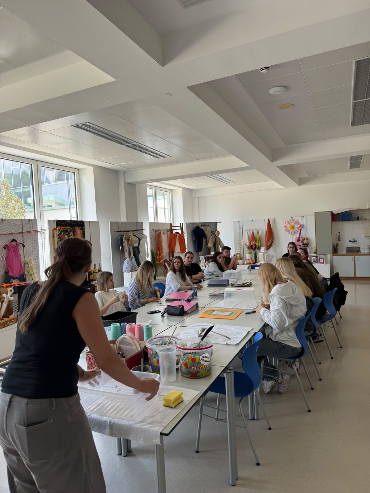 Die 3.PA beim Workshop "Papierschöpfen" Heller Klassenraum mit mehreren Personen an Tischen, die gemeinsam an bunten Materialien arbeiten; im Vordergrund sitzt eine Person, weitere Personen stehen oder sitzen im Hintergrund.