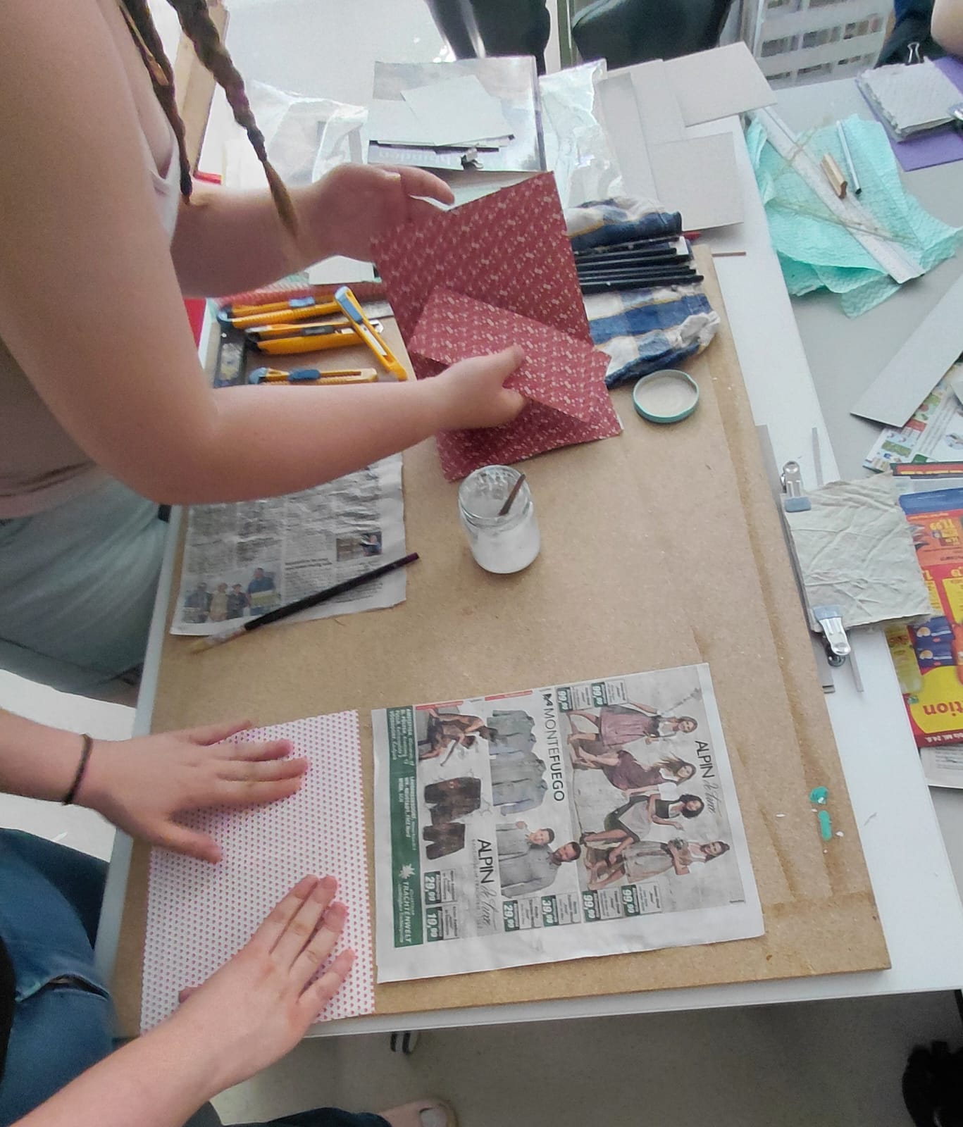 Workshop "Buchbinden" Ein Tisch mit Schneideunterlage, Zeitungspapier und Stanleymessern, sowie weitern Untensilien. Vier Hände halten Papiere.