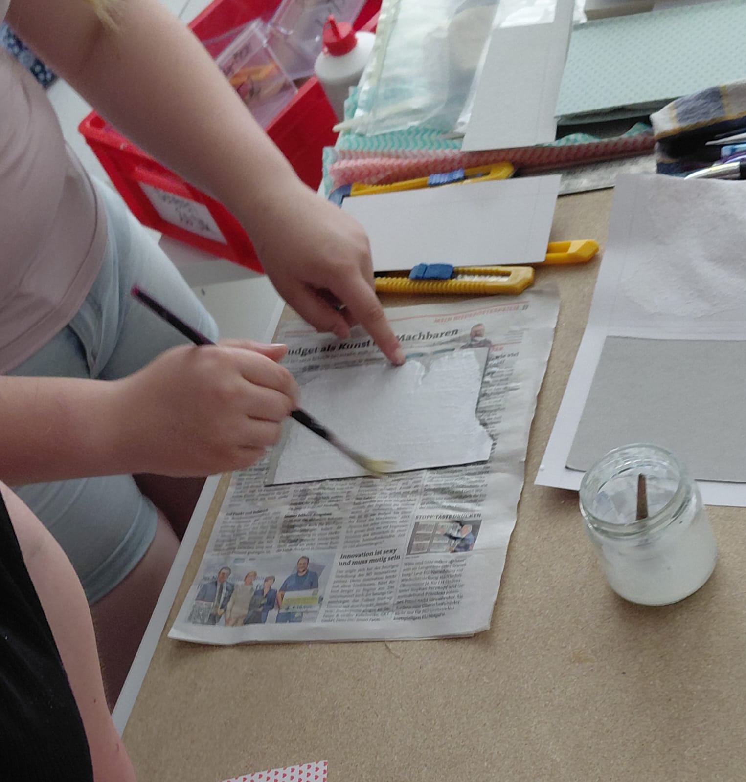 Workshop "Buchbinden" Zwei Hände, die auf einer mit Zeitung belegten Arbeitsplatte ein kleineres Stück Papier mit weißem Leim einpinseln und das Papier mit einem Finger halten. Daneben liegen Schneidewerkzeuge und andere Utensilien.