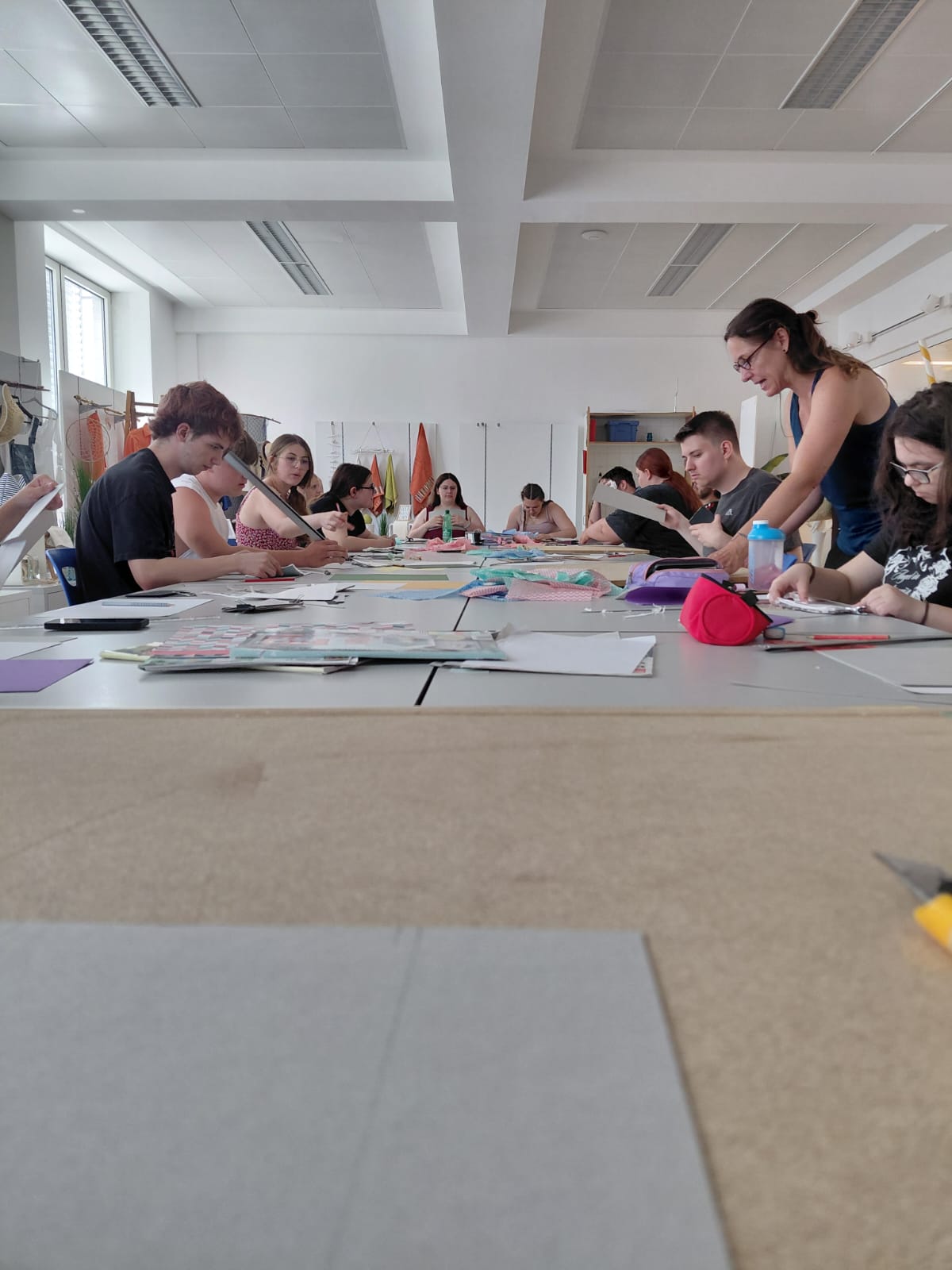 Workshop "Buchbinden" - 1. PA Blick über eine Arbeitsfläche in eine Klasse. Die Tische sind zusammengestellt, rundum sitzen Schüerlinnen und Schüler, die mit Papier beschäftigt sind. Eine Trainerin steht zwischen den Jugendlichen.