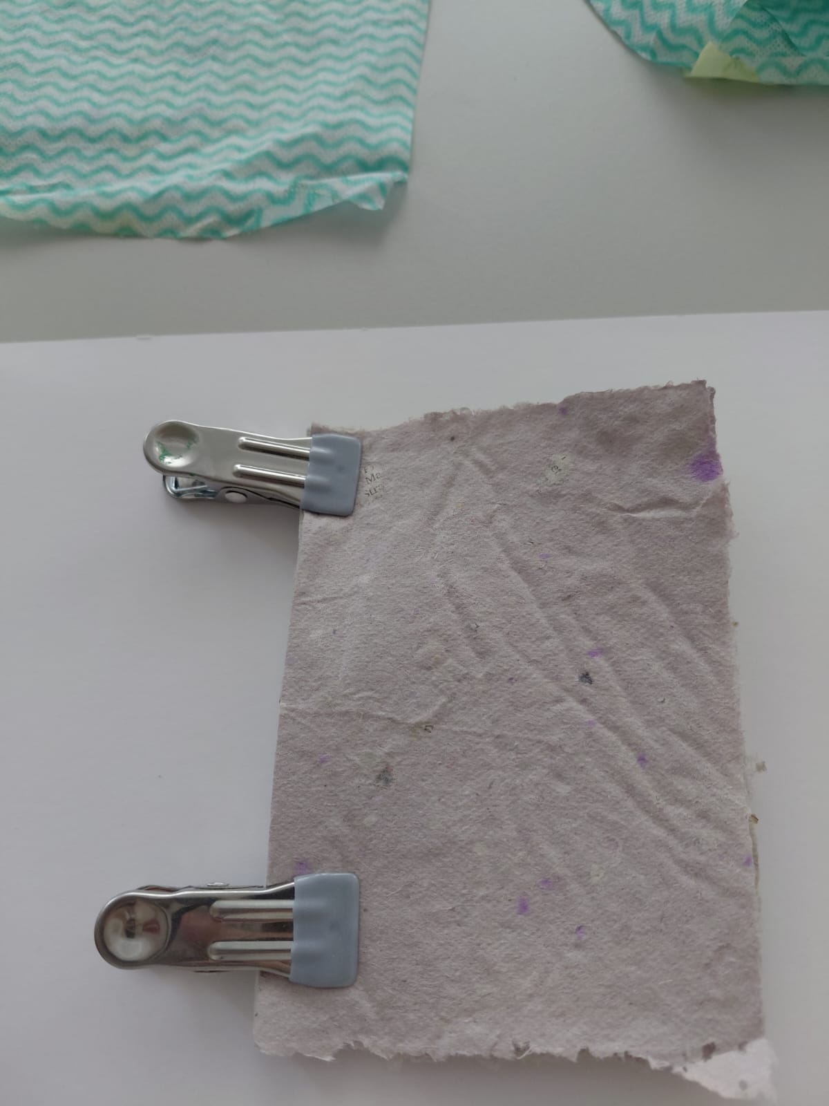 Workshop "Buchbinden" Büttenpapier mit zwei Klammern zusammengehalten auf einem weißen Blatt, im Hintergrund Küchentücher mit Zackenmuster.