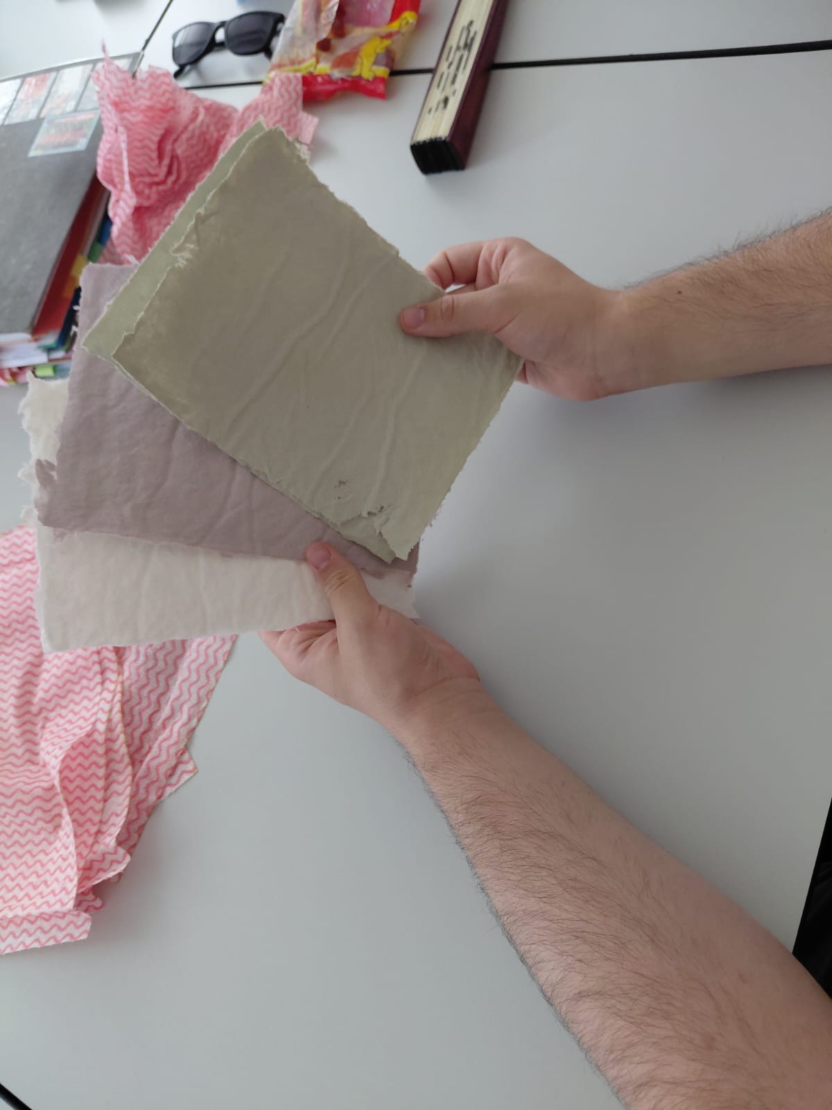 Workshop "Buchbinden" Zwei Hände halten drei Blätter Büttenpapier aufgefächert. Jedes Blatt hat eine unterschiedliche Farbe: weiß, rosa, graubraun.