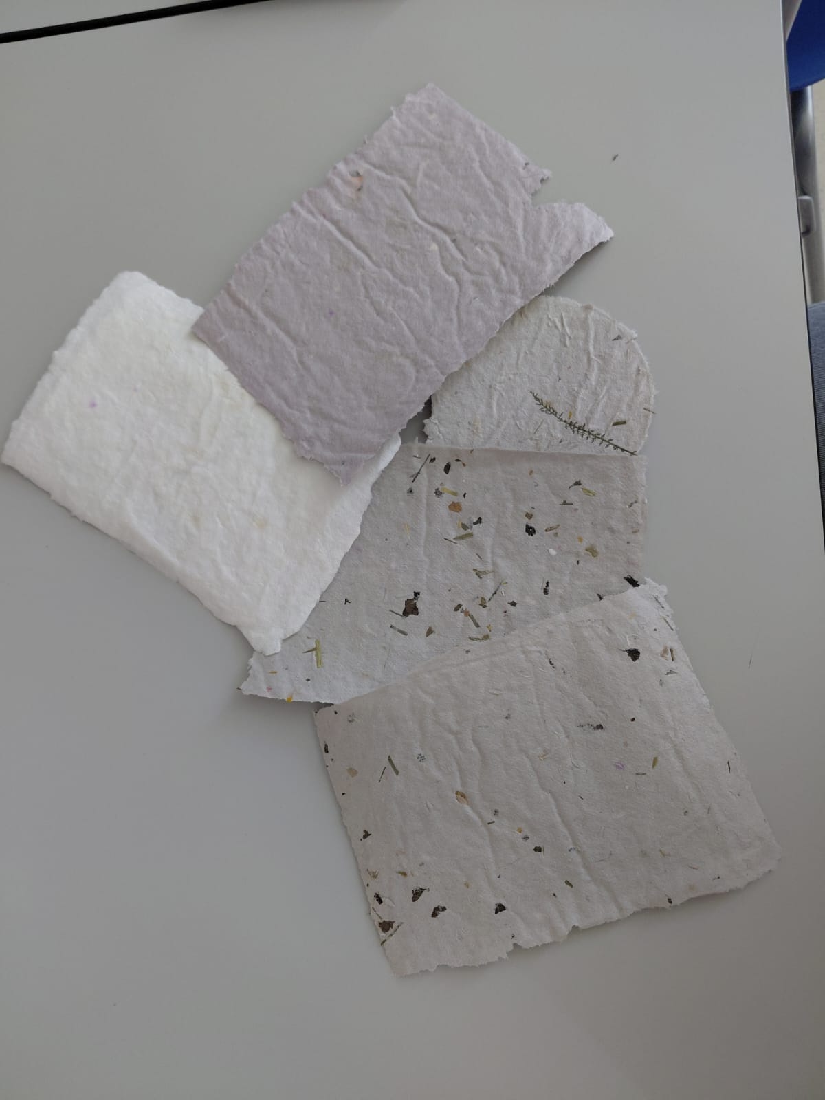 Workshop "Buchbinden" Mehrere Blätter Büttenpapier auf einem Tisch. Sie haben unterschiedliche Farbtöne und teilweise Blüten und Blätter eingearbeitet.