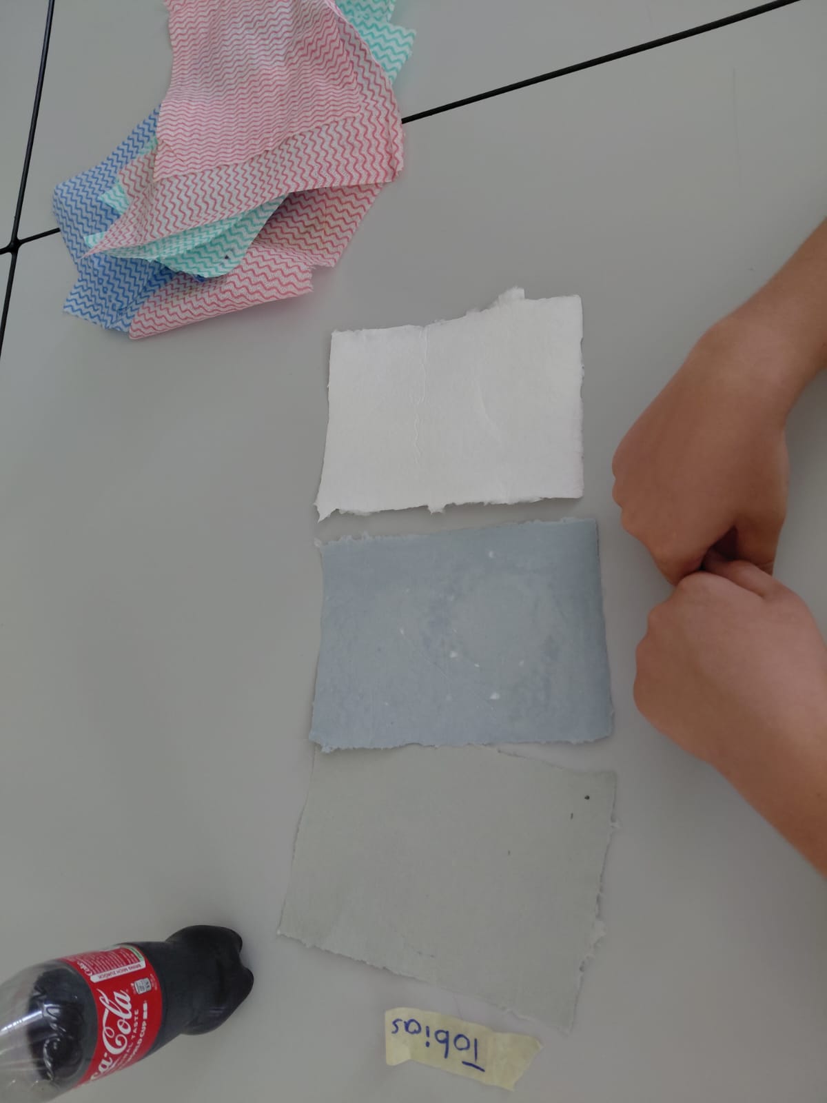 Workshop "Buchbinden" Zwei locker geschlossen gehaltene Hände über einem Tisch mit drei Blatt Büttenpapier in grau, blau und weiß, daneben ein Sticker mit dem Namen "Tobias", eine Cola Flasche und mehrere Küchentücher.