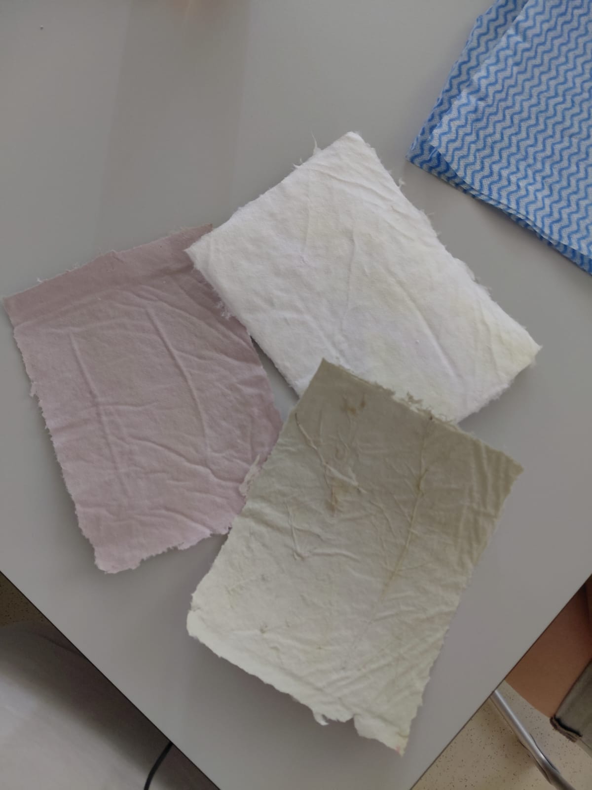 Workshop "Buchbinden" Drei Blatt Büttenpapier in beige, rosa und Weiß und ein blau gemustertes Küchentuch auf einem Tisch.