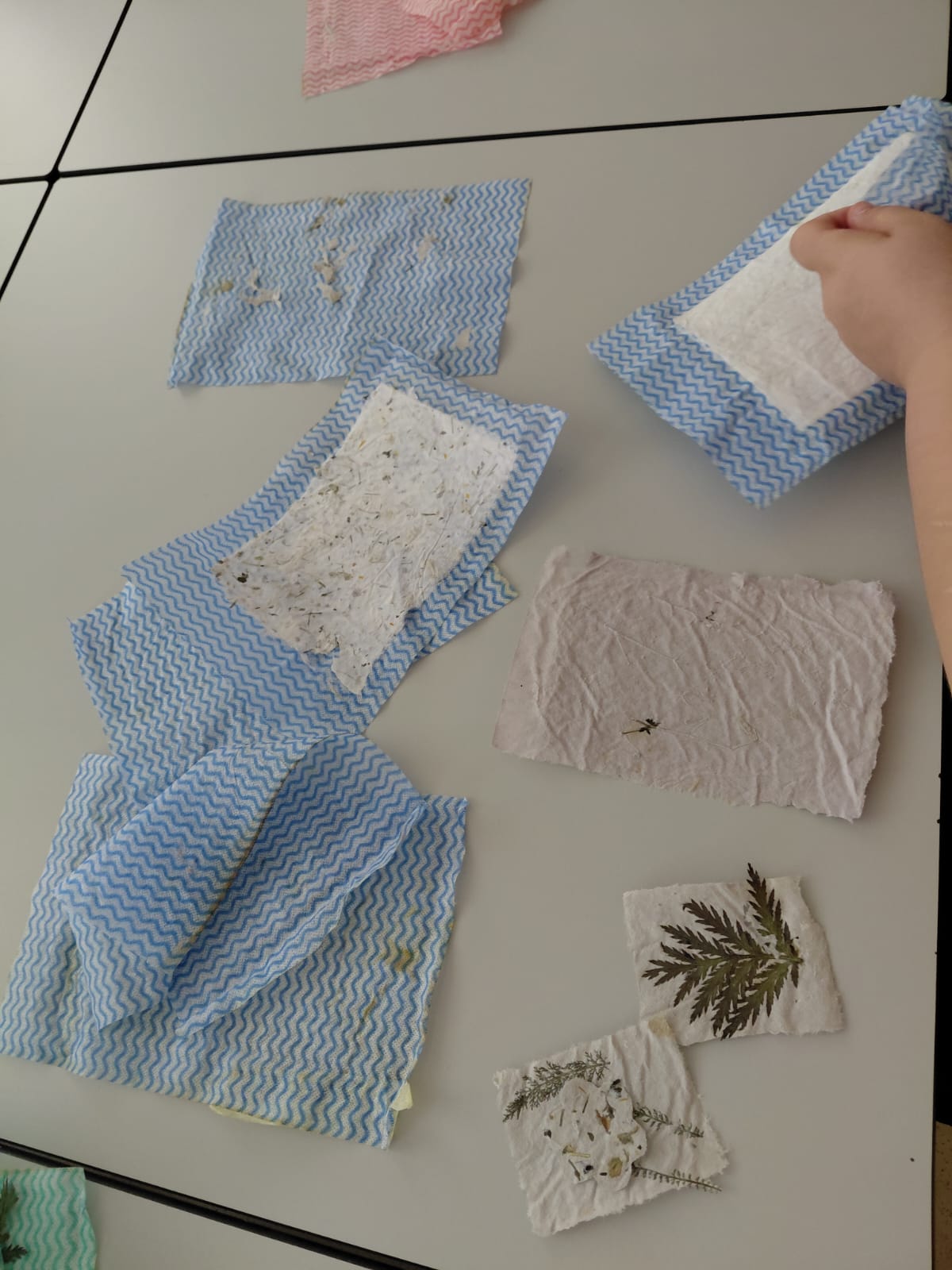 Workshop "Buchbinden" Ein Tisch mit blau gemusterten Küchentüchern, teilweise mit einem Blatt Büttenpapier darauf. Daneben zwei kleine Stücke Büttenpapier mit eingearbeiteten Pflanzen, ein größeres Stück Papier und eine Hand, die ein Küchentuch mit Papier hält.