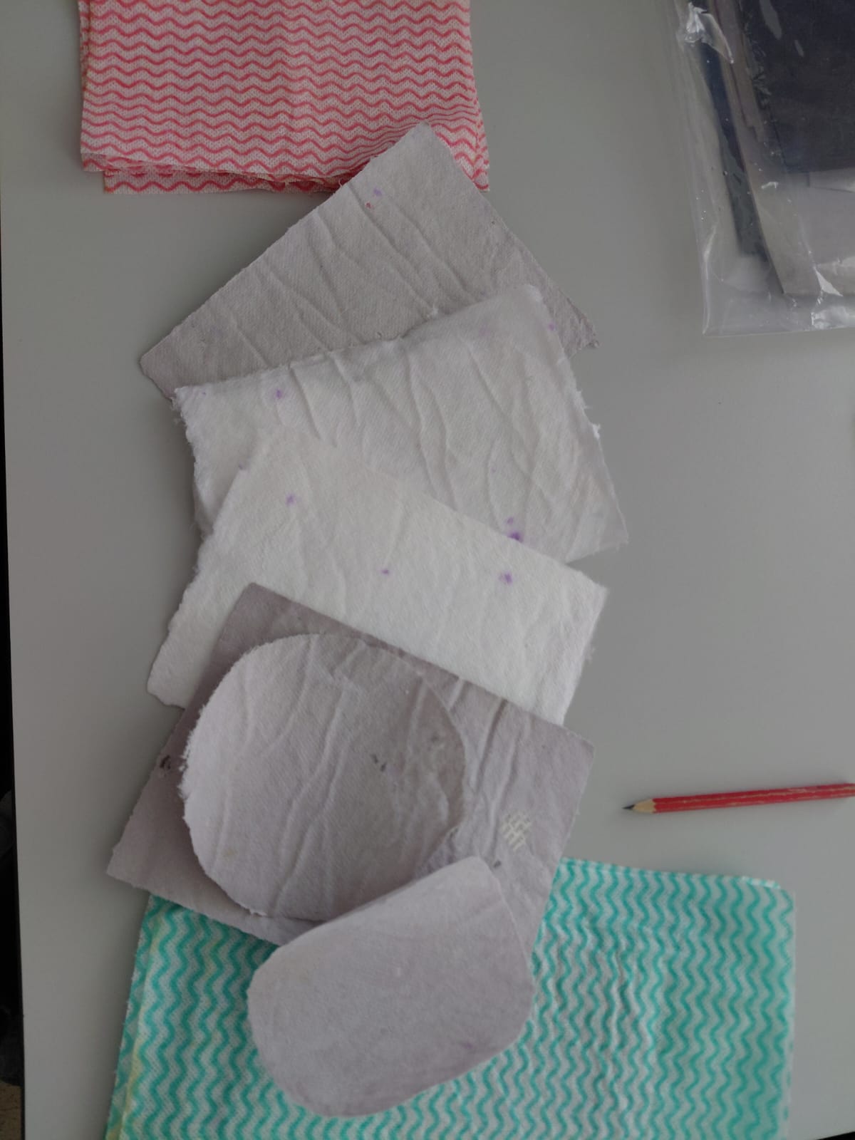 Workshop "Buchbinden" Auf einem Tisch lieben aufgefächert mehrere Blätter Büttenapier in unterschiedlichen weiß und lila-Tönen. Daneben ein Bleistift und rote und grüne Küchentücher.