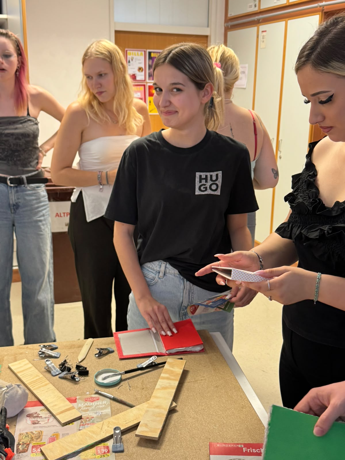 Workshop "Buchbinden" Mehrere junge Frauen stehen um einen Tisch mit verschiedenen Utensilien, wie Pinseln, Klammern und Papier. Eine davon hält den Buchdeckel für ein kleines Buch, eine weitere sieht mit einem beginnenden Lächeln in die Kamera.
