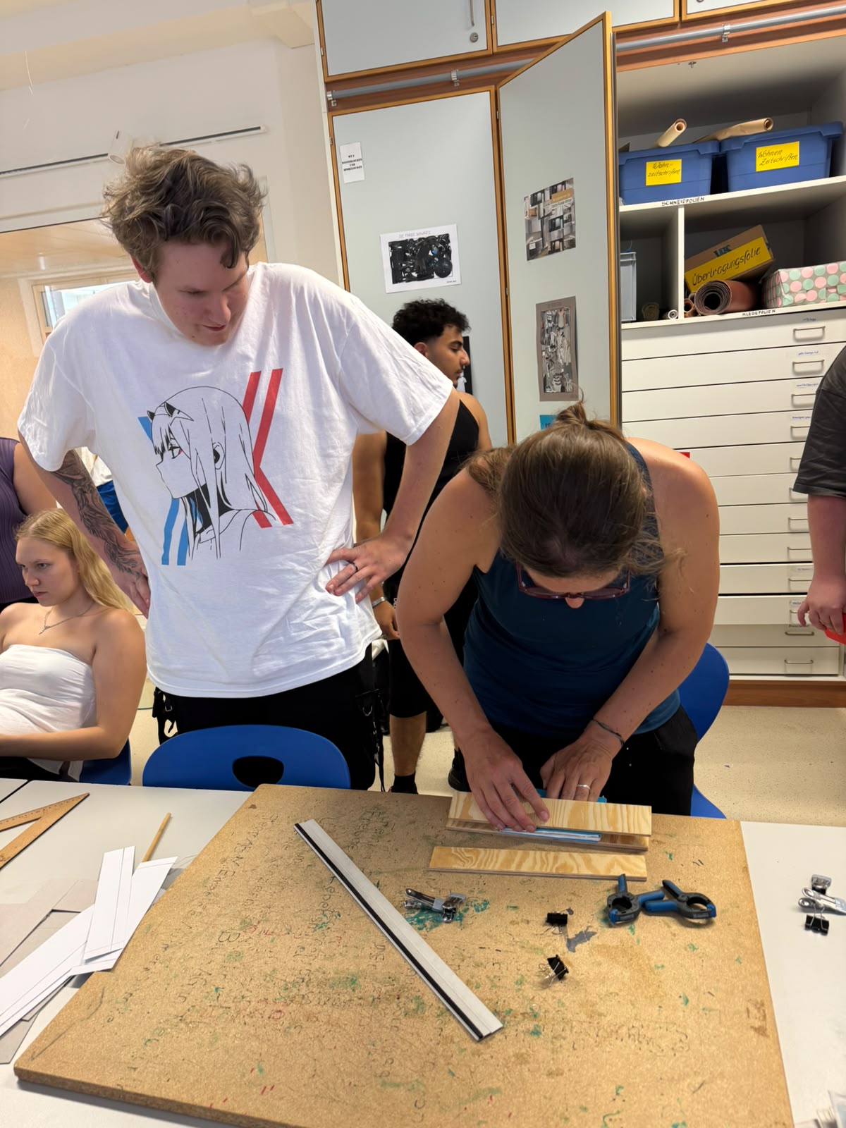Workshop "Buchbinden" Eine junge Frau beugt sich über die Arbeitsfläche mit Schneideunterlage, Papier und Lineal. Ein junger Mann steht daneben und sieht zu. Im Hintergrund sind weitere Jugendliche und ein offener Schrank mit vielen breiten niedrigen Schubladen zu sehen.